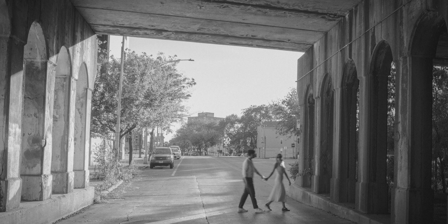 Black and white photograph of a covered walkway with arched columns and silhouetted figures walking.