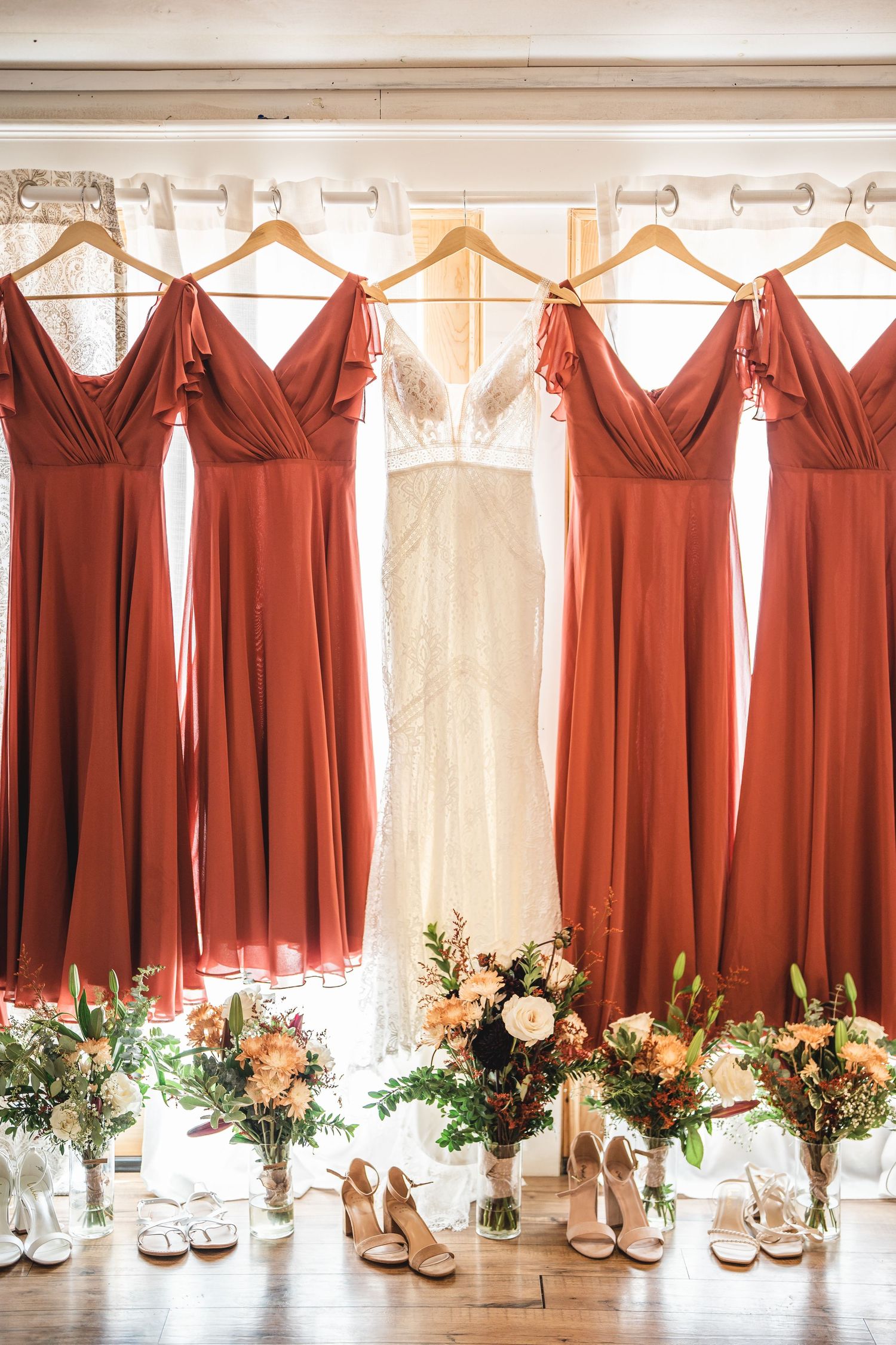 Row of rust-colored bridesmaid dresses hanging by windows with floral bouquets below.
