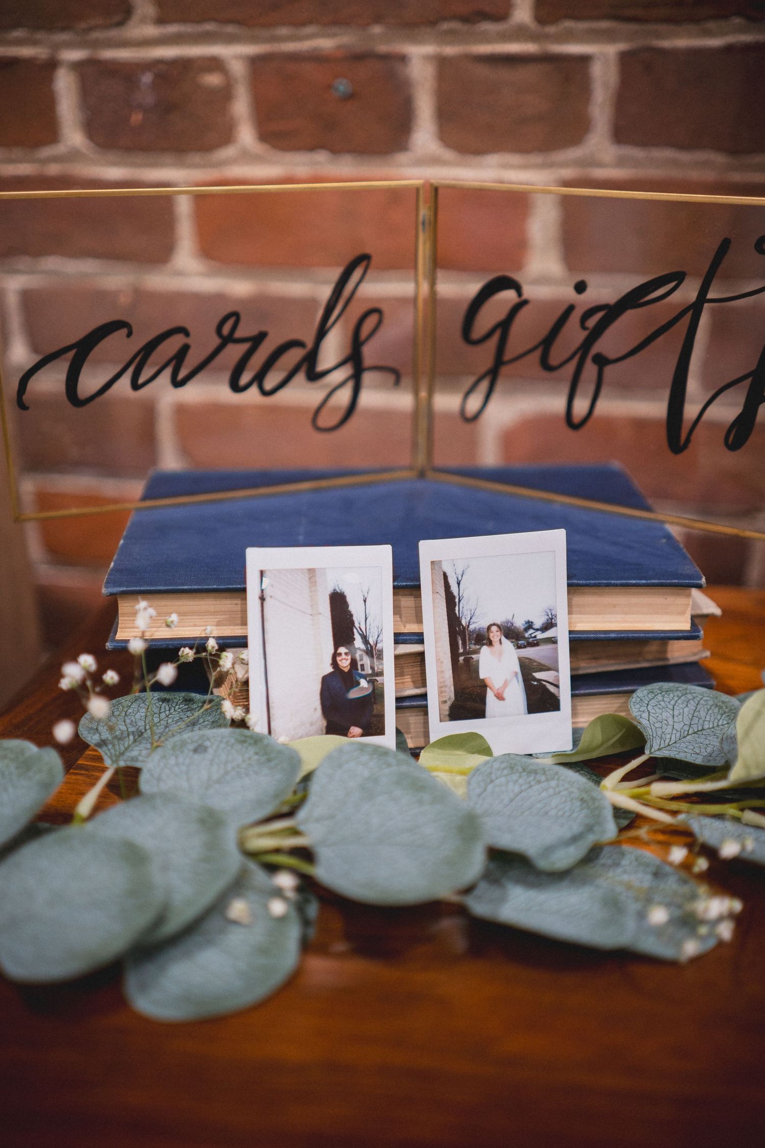 Wedding cards and gifts display with eucalyptus garland and polaroid photos against a brick wall.