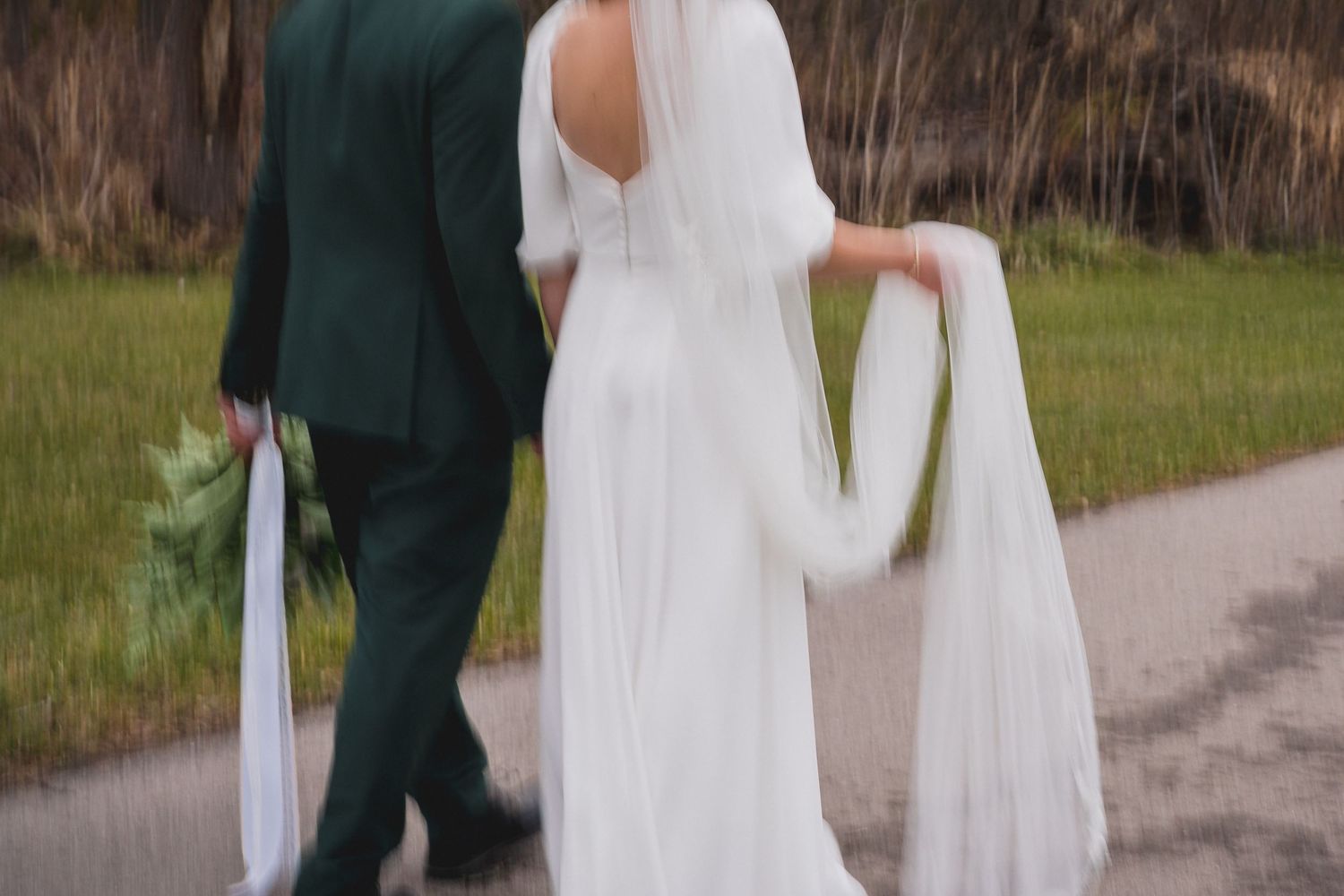 A couple walking together on a path, with flowing white wedding dress and emerald green suit.