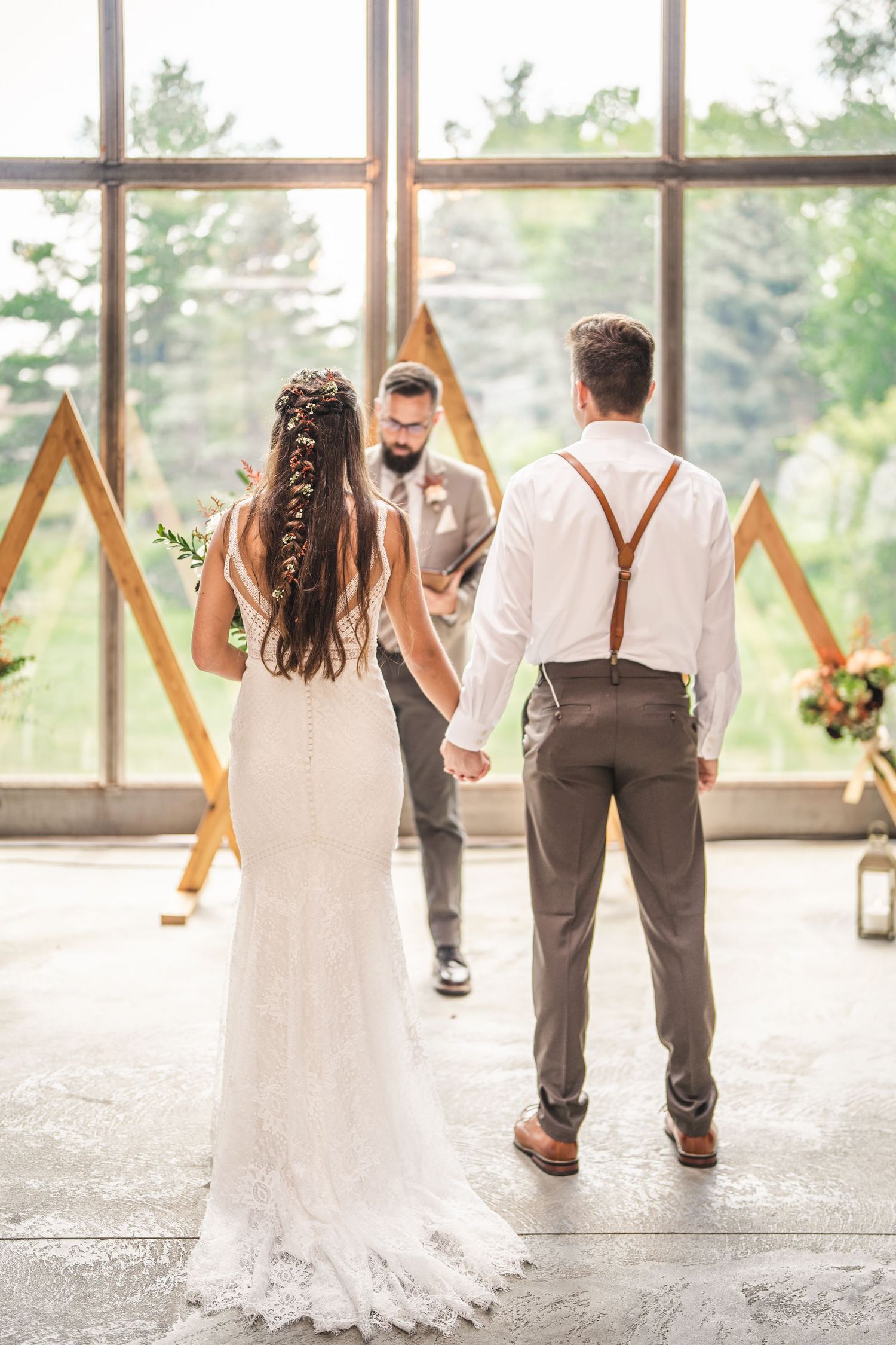 Couple stands before geometric wooden arch during wedding ceremony with natural light streaming through windows.