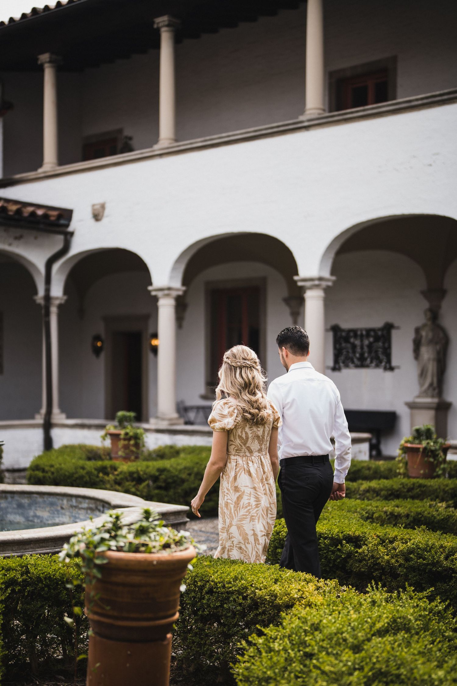 Couple walking through a Spanish colonial courtyard with white columns and green hedges.