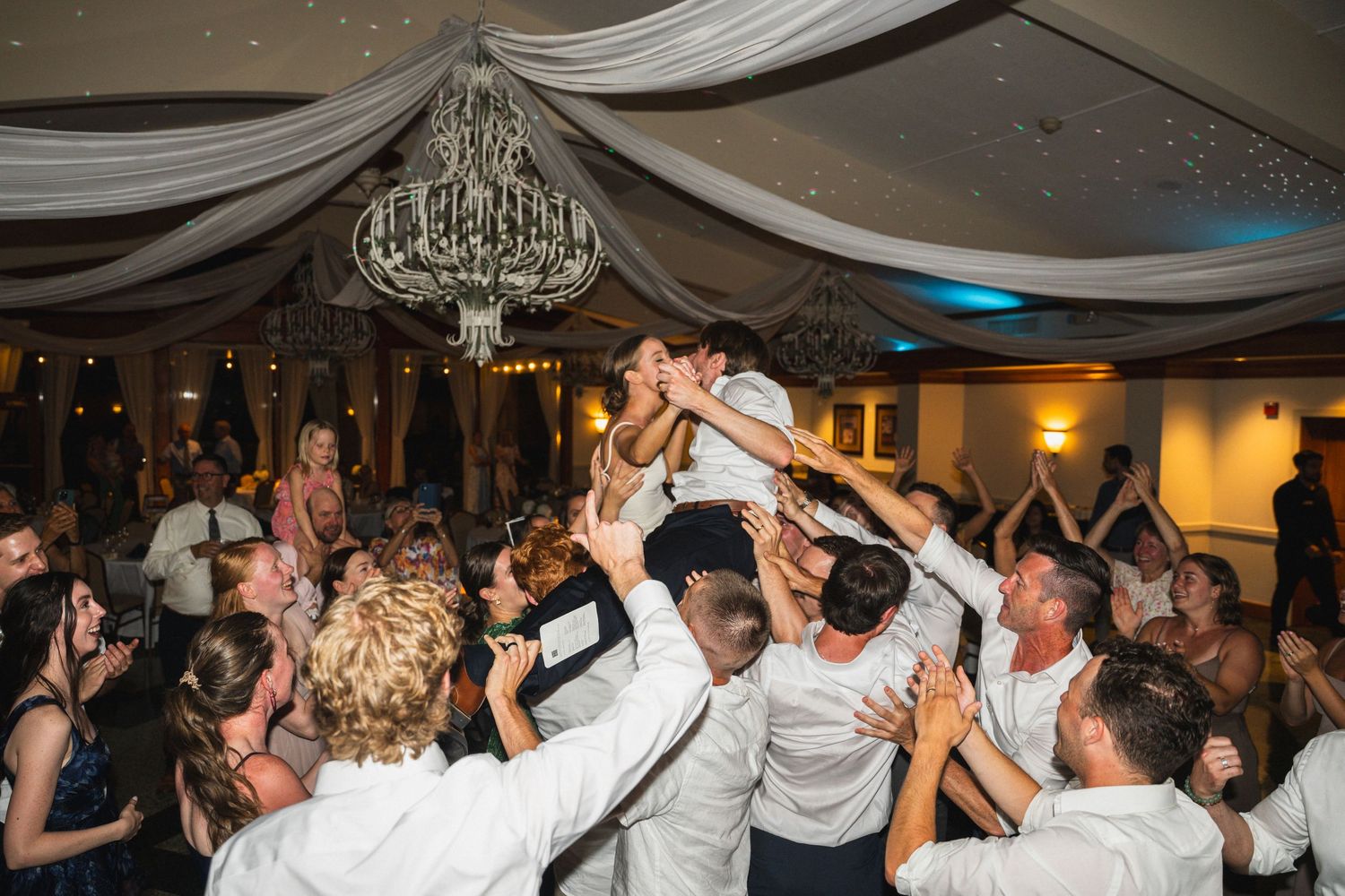 Excited wedding guests lift the bride during a lively reception dance floor celebration.