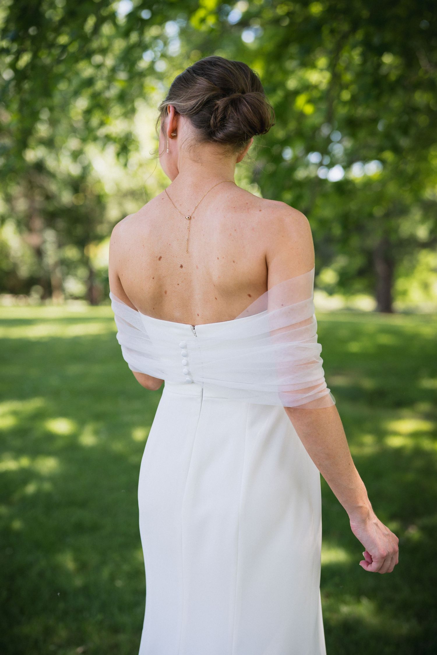 Bride in white off-shoulder gown with pink wrap showing elegant back detail in lush green park setting.
