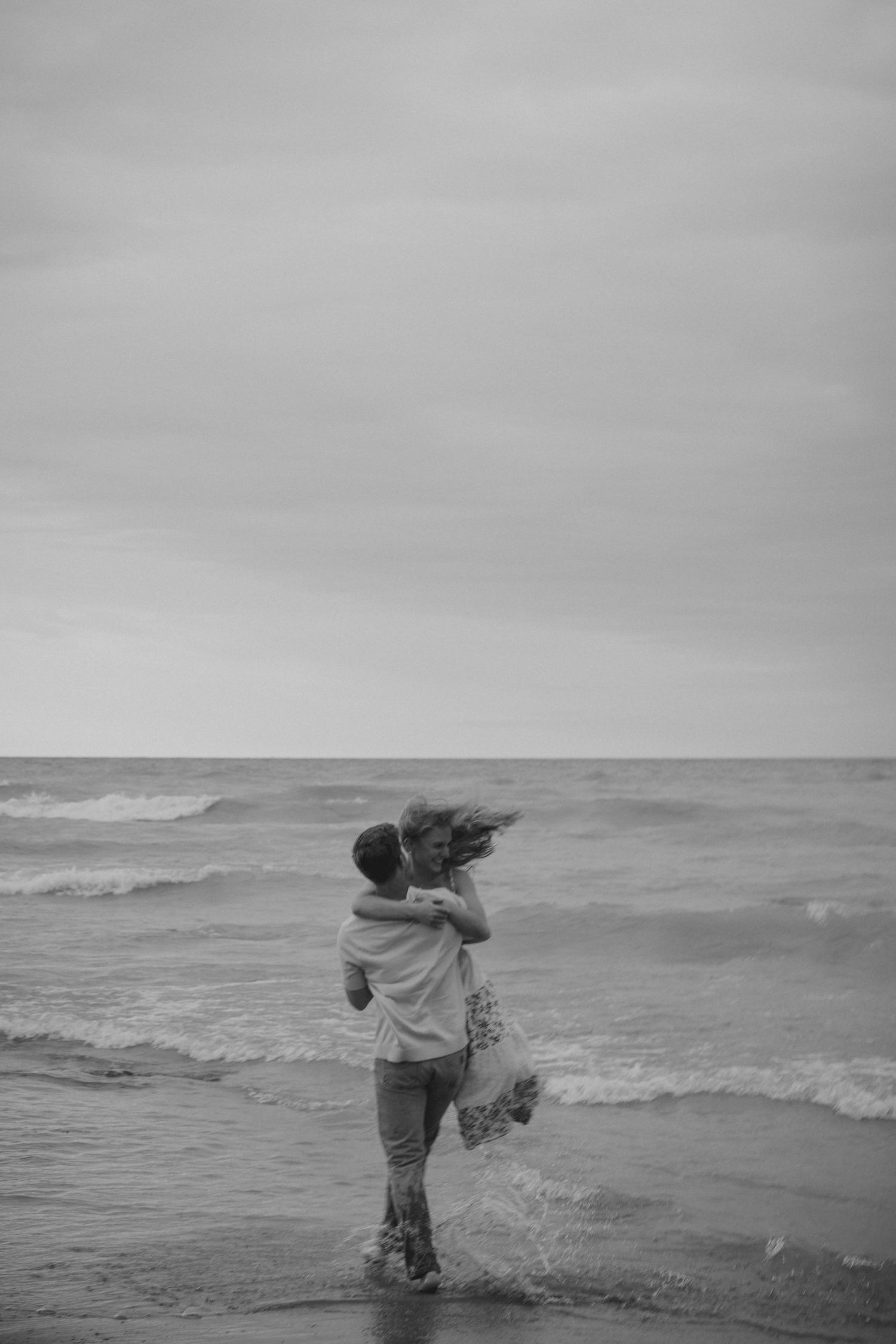 Black and white photo of romantic couple embracing in ocean waves on a cloudy beach day.