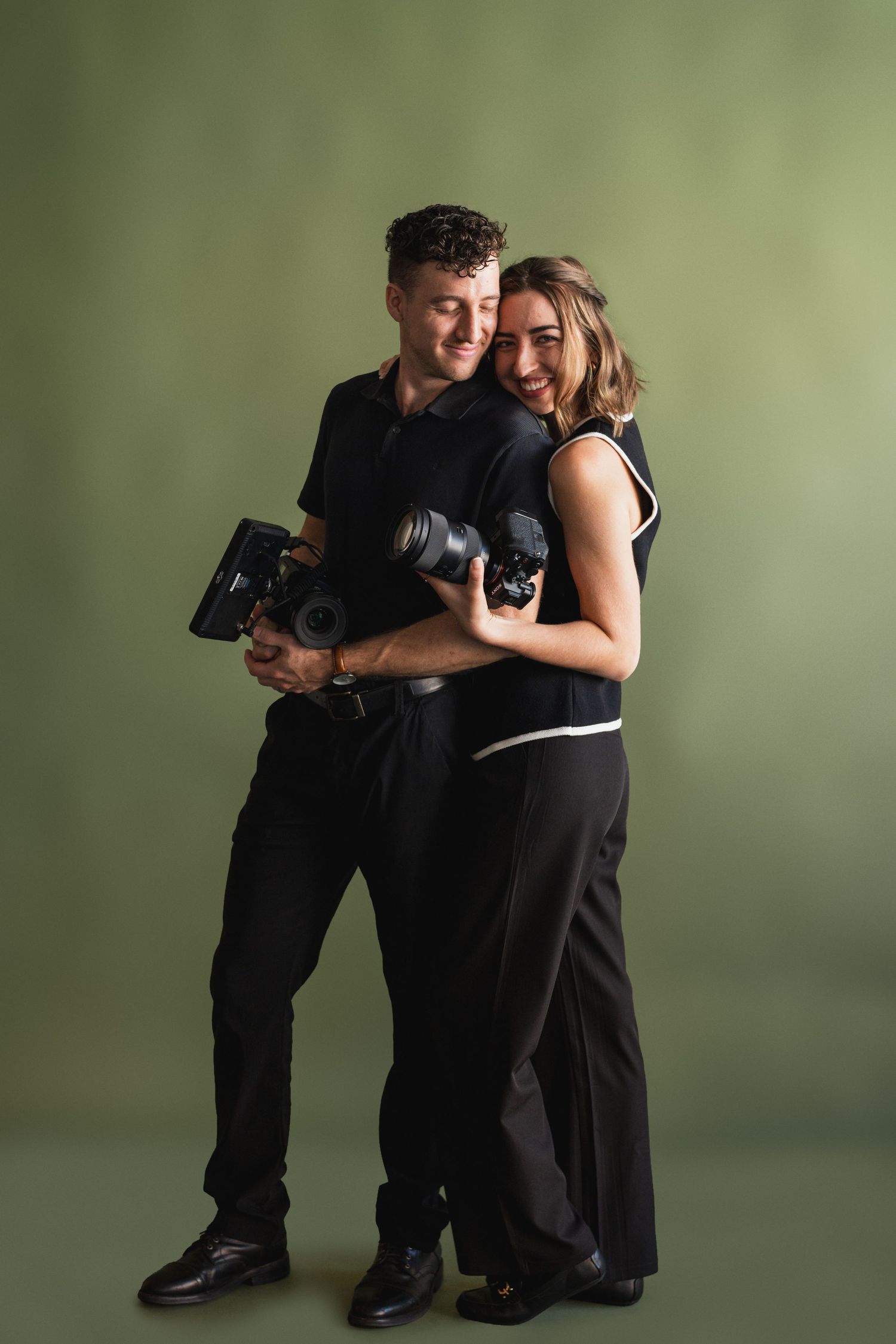 Photography and filmmaking duo poses with cameras against green studio backdrop.