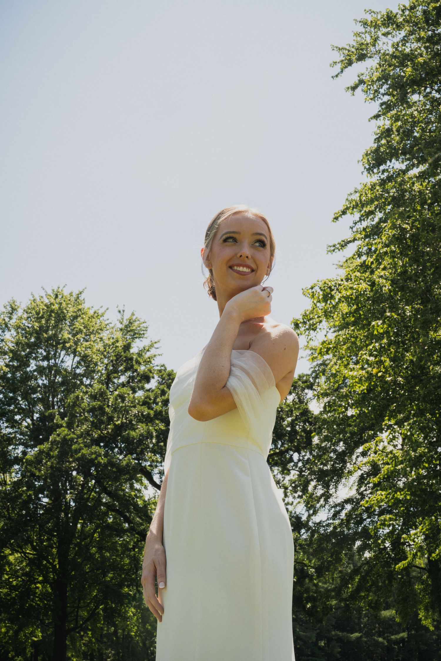 Smiling bride in white strapless gown looks up joyfully against blue sky framed by green trees.