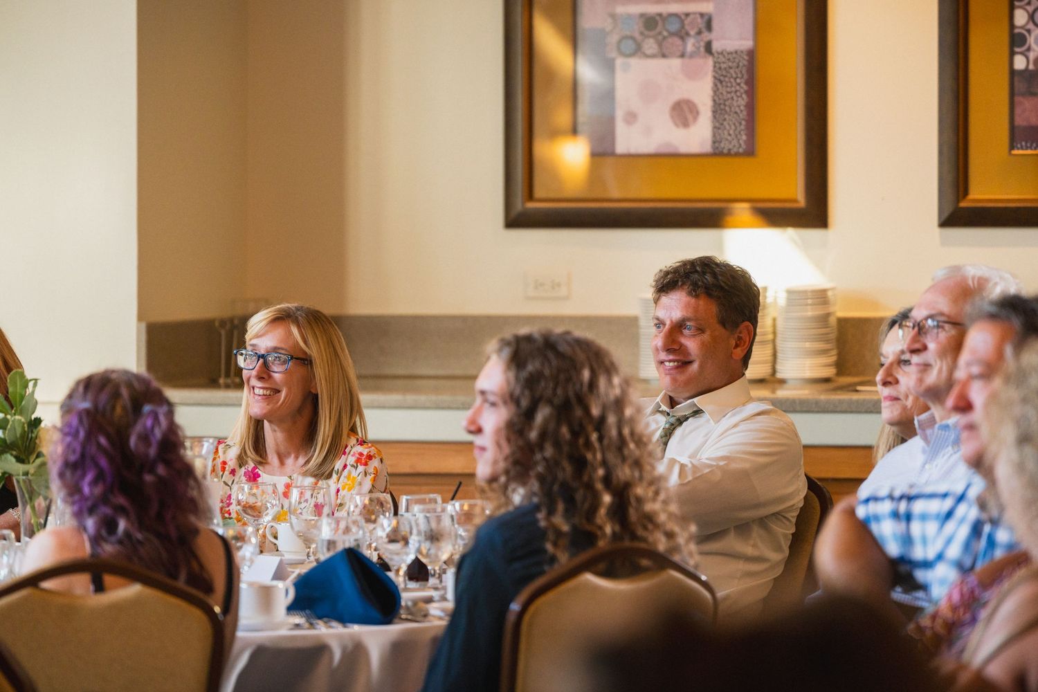 Guests laughing and smiling at a wedding reception dinner table with wine glasses.