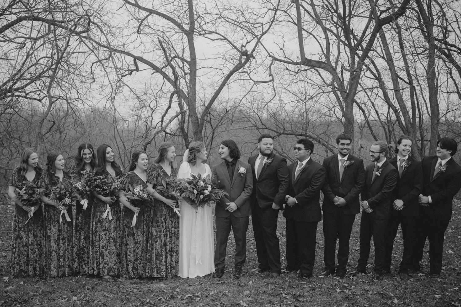 Wedding party poses outdoors in winter with bridesmaids in dark dresses and groomsmen in suits.