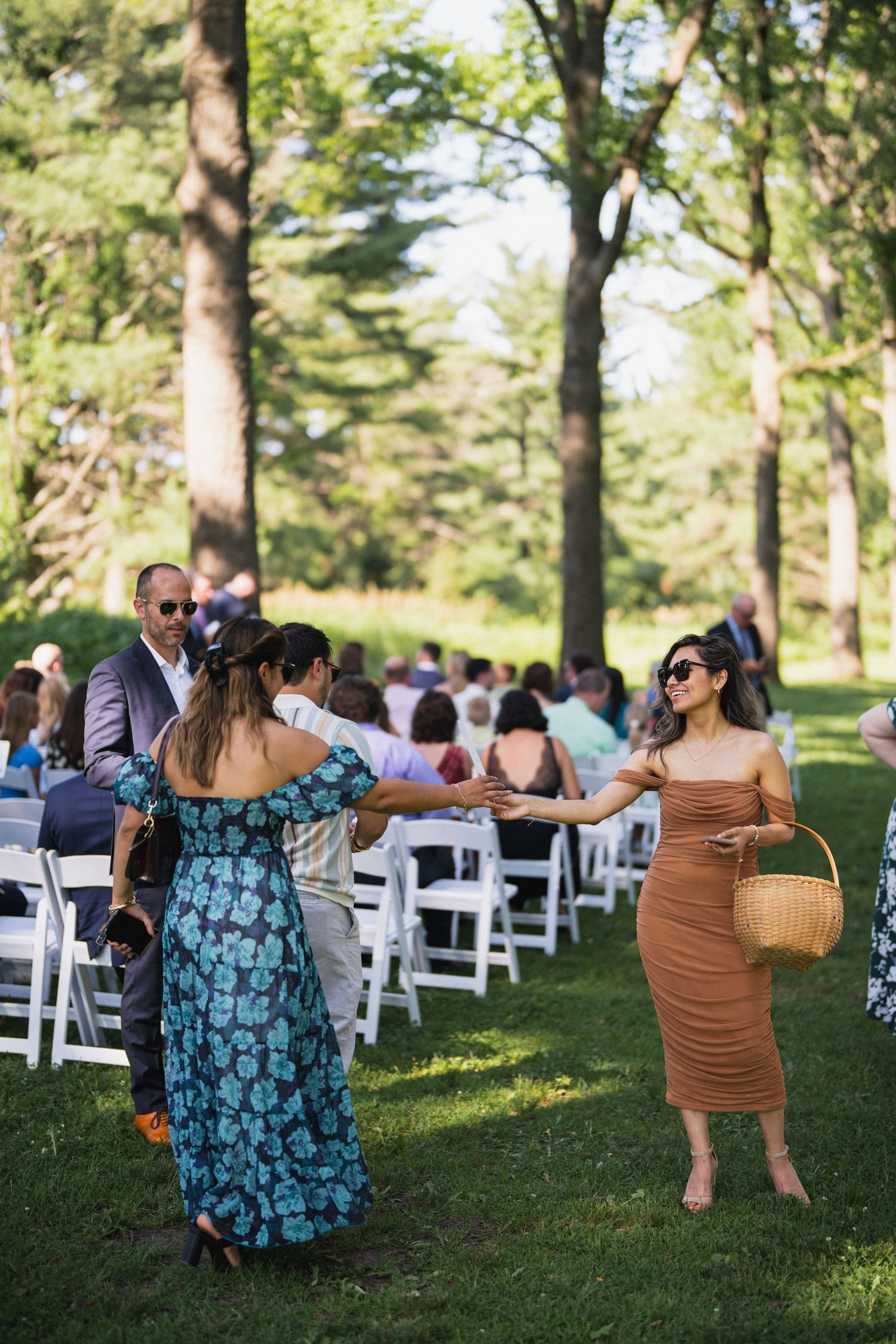 Two women greeting each other at an outdoor wedding ceremony with white chairs and lush green trees.