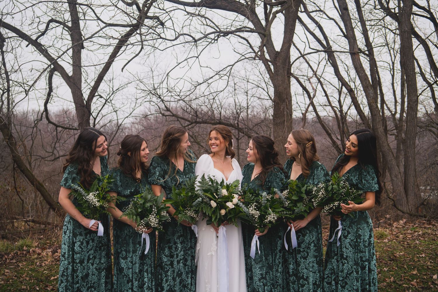Bridesmaids in emerald velvet dresses stand with bride holding greenery bouquets against bare trees.