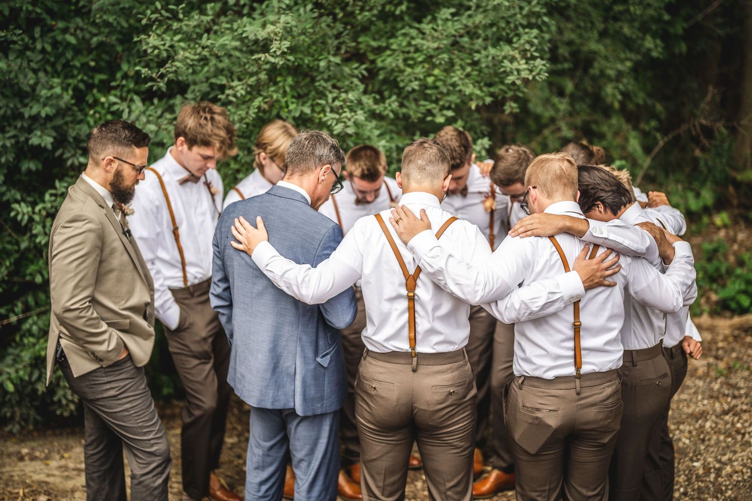 Group wearing suspenders and suits huddle together outdoors with greenery in background.