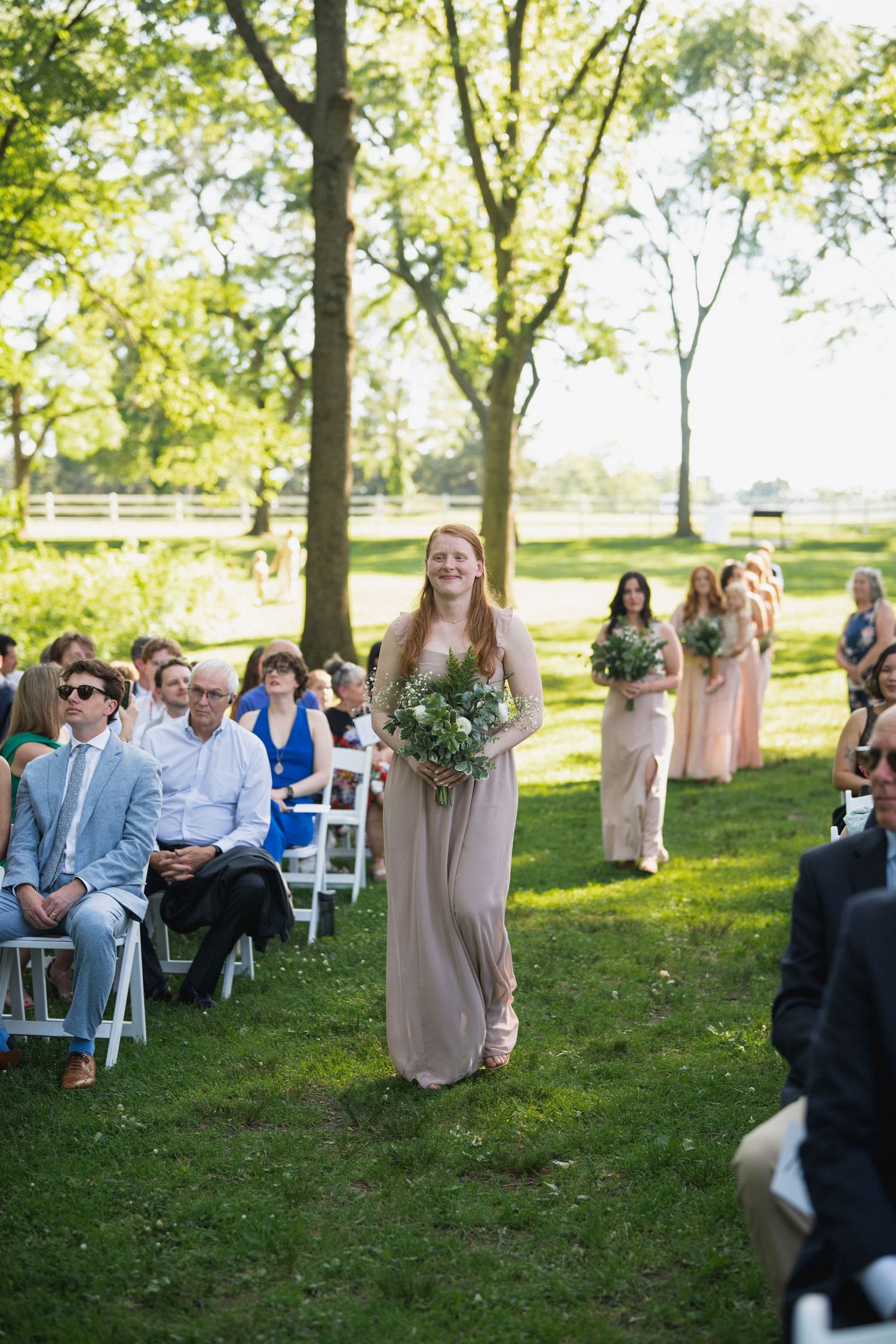 Smiling bridesmaid in a mauve gown carrying a white floral bouquet walks down the outdoor wedding aisle.