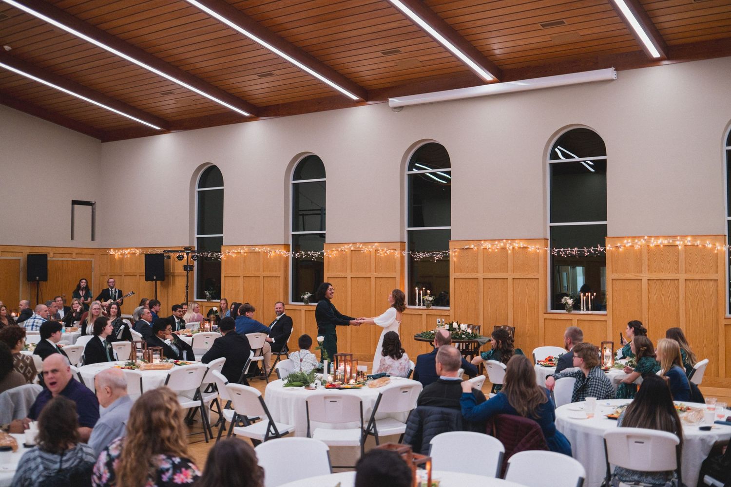 Wedding reception venue with arched windows and string lights as guests sit at round tables.