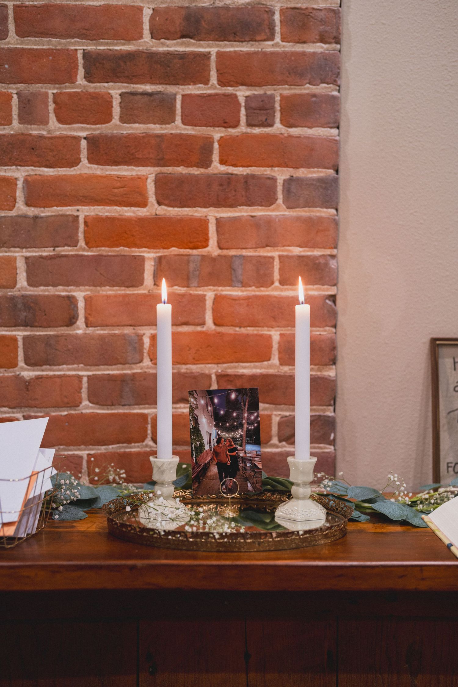 White candles and decorative greenery arranged on a wooden table against exposed brick wall.
