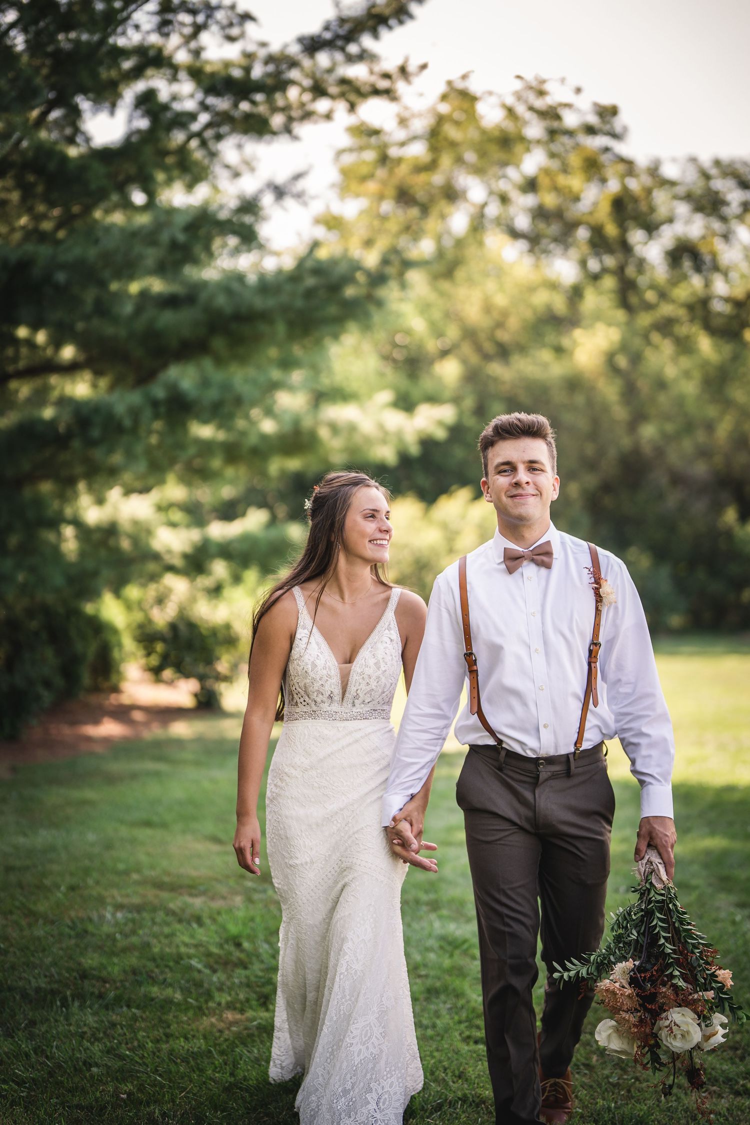 Newlyweds stroll through a sunlit garden holding hands and bouquet.