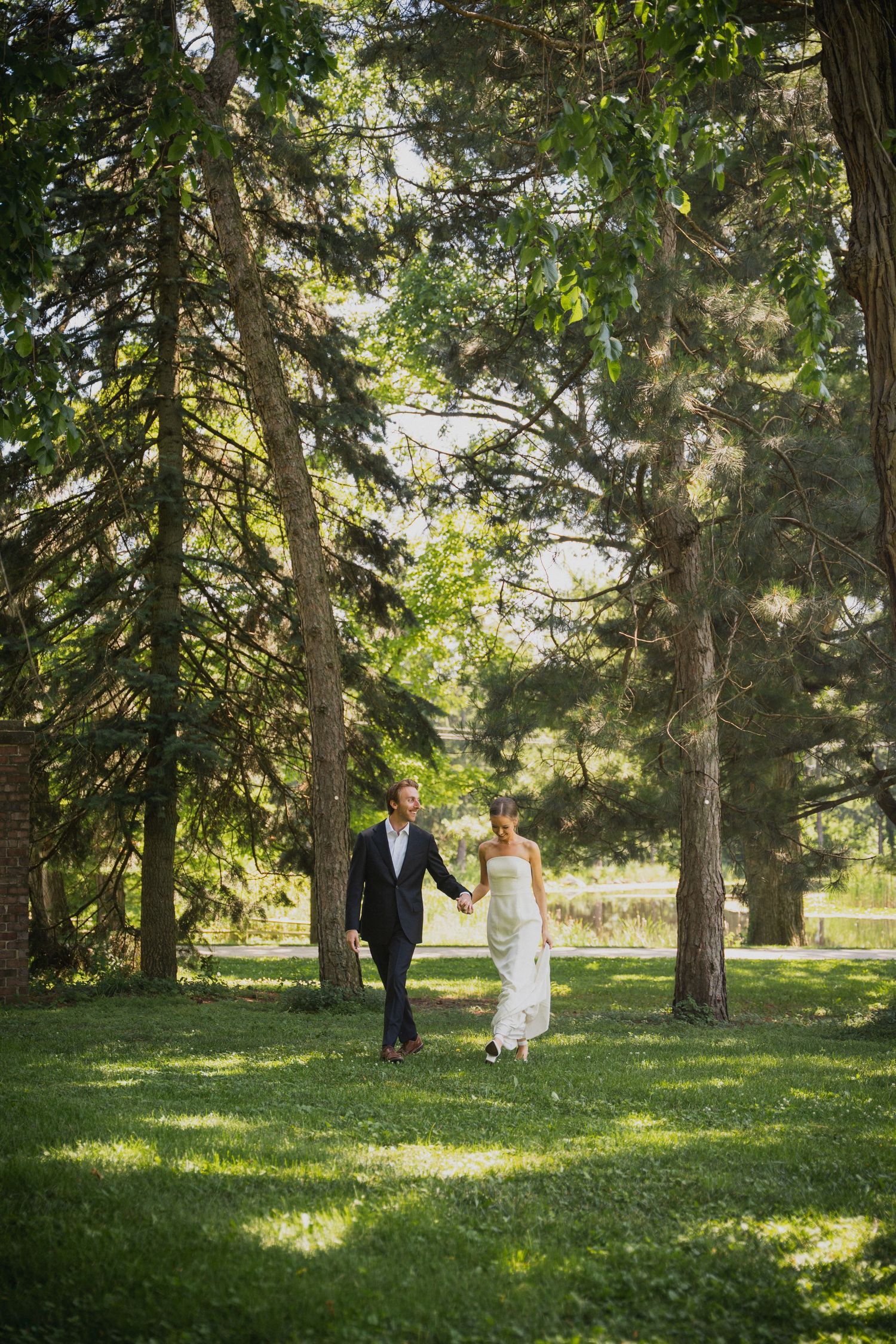 A couple walking hand in hand through a sunlit forest in formal attire.