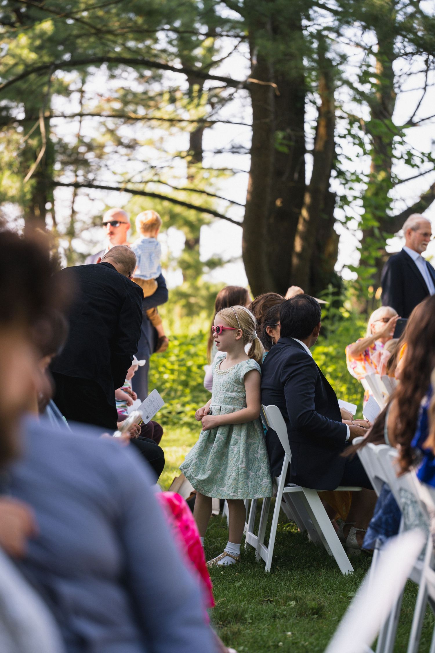 Young girl in a mint dress and pink sunglasses stands among seated wedding guests at an outdoor ceremony.