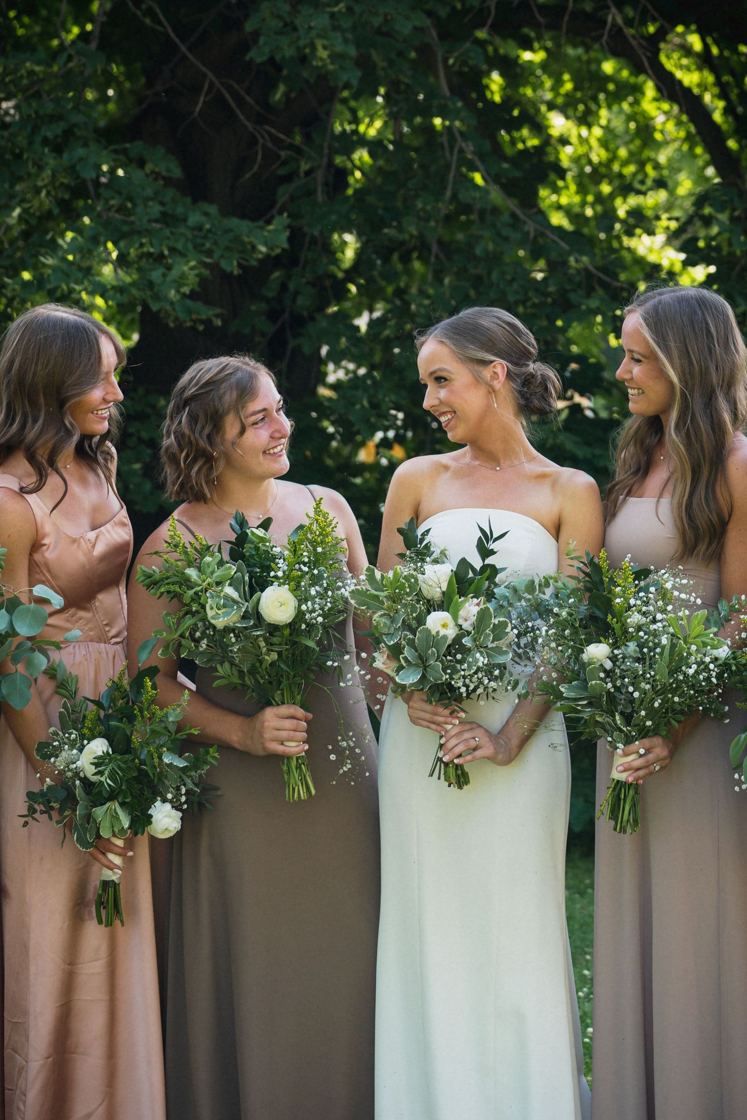 Bride in strapless white gown poses with three bridesmaids in dusty rose dresses holding greenery bouquets outdoors.