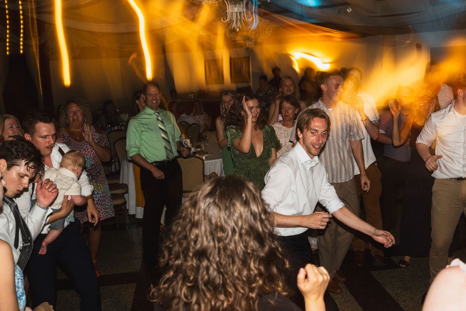 Lively wedding reception dance floor with guests dancing and celebrating under warm golden lighting.
