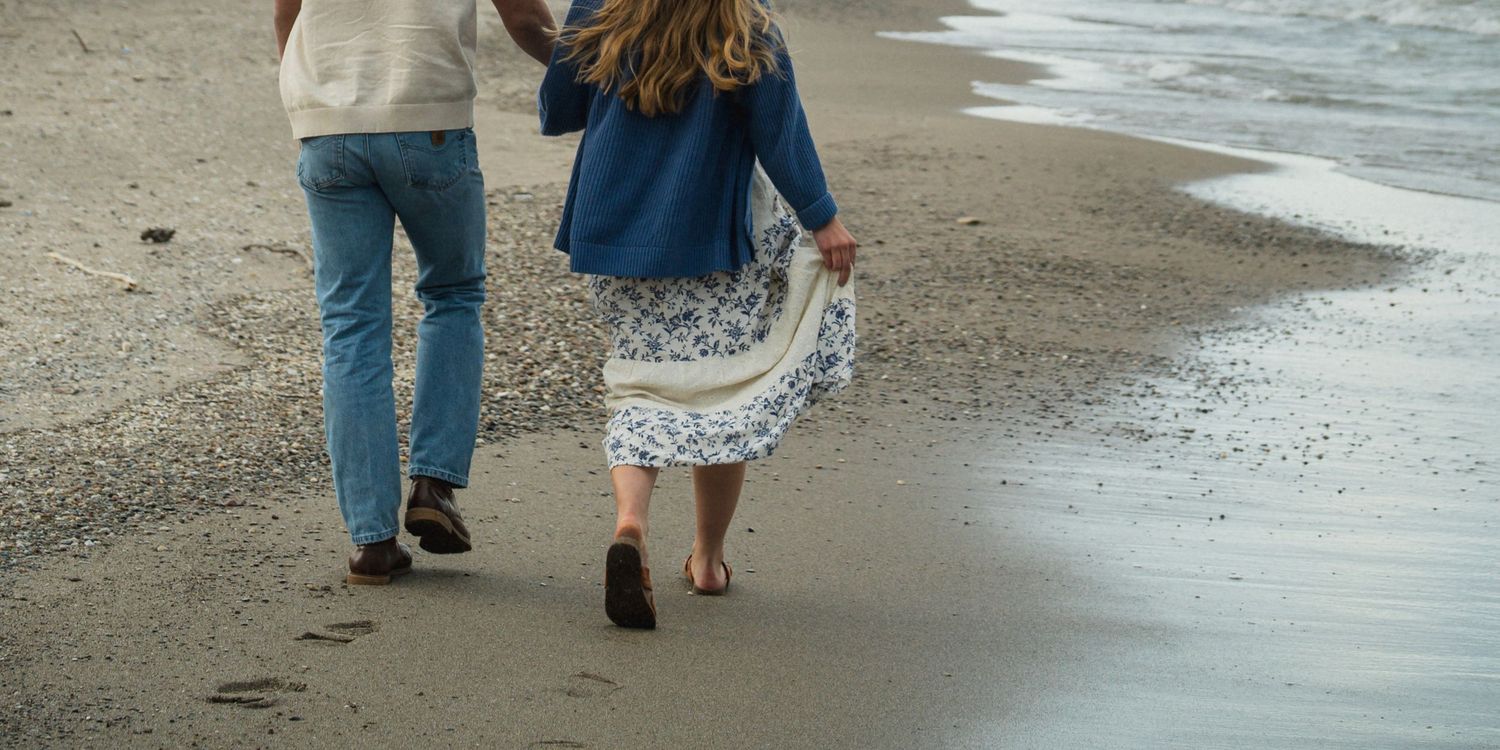 Couple holding hands walking barefoot on a sandy beach shore, woman in floral skirt and blue jacket.