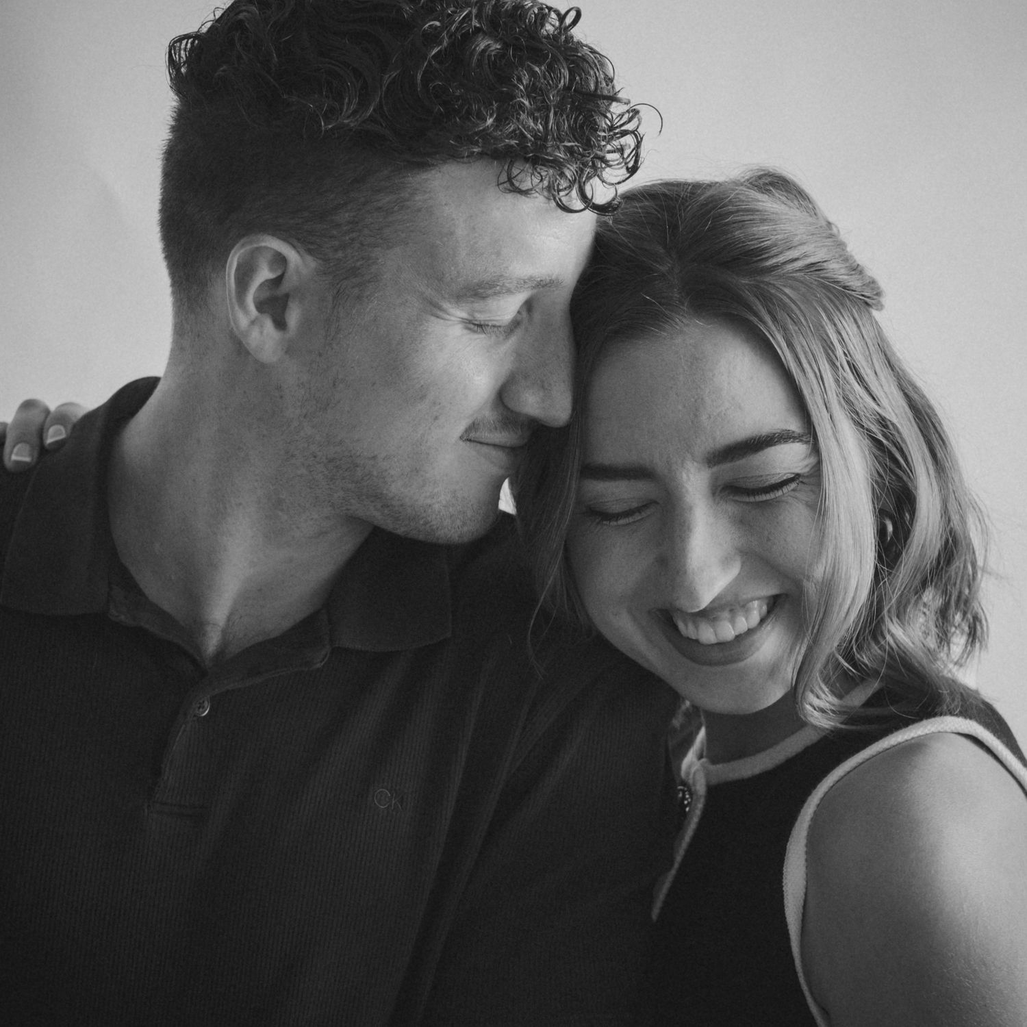 Intimate black and white portrait of couple sharing tender moment against plain background.