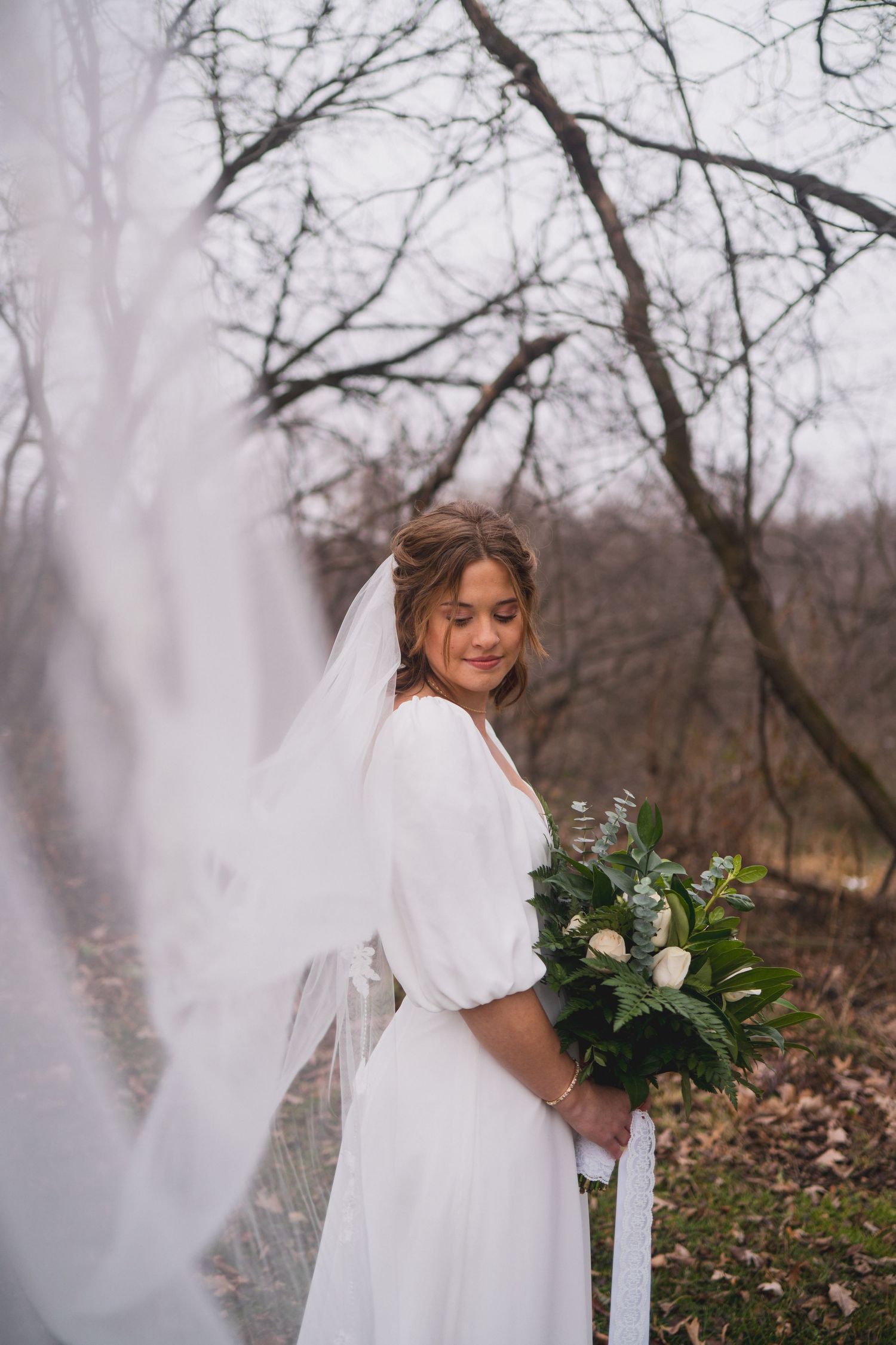Ethereal bridal portrait with flowing veil and white bouquet in a winter forest setting.