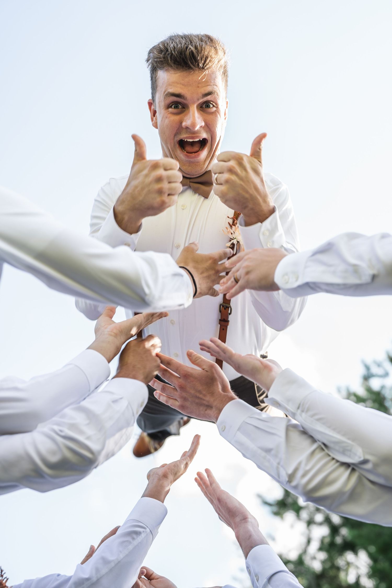 Groom in white shirt and bowtie being lifted by groomsmen's hands reaching from below.