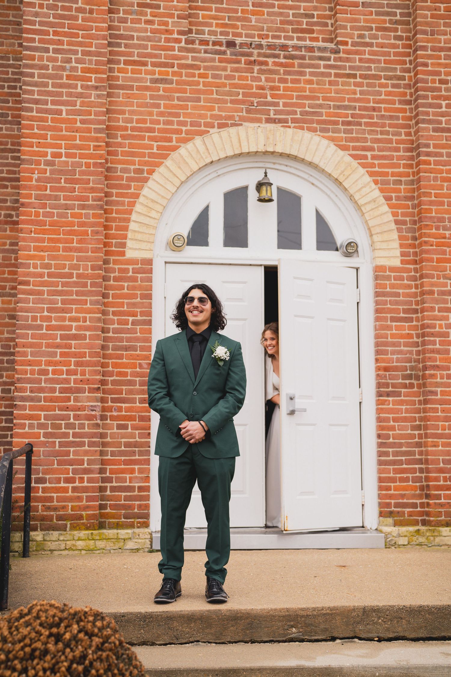 Person in green suit poses playfully outside brick church building with white arched doors.
