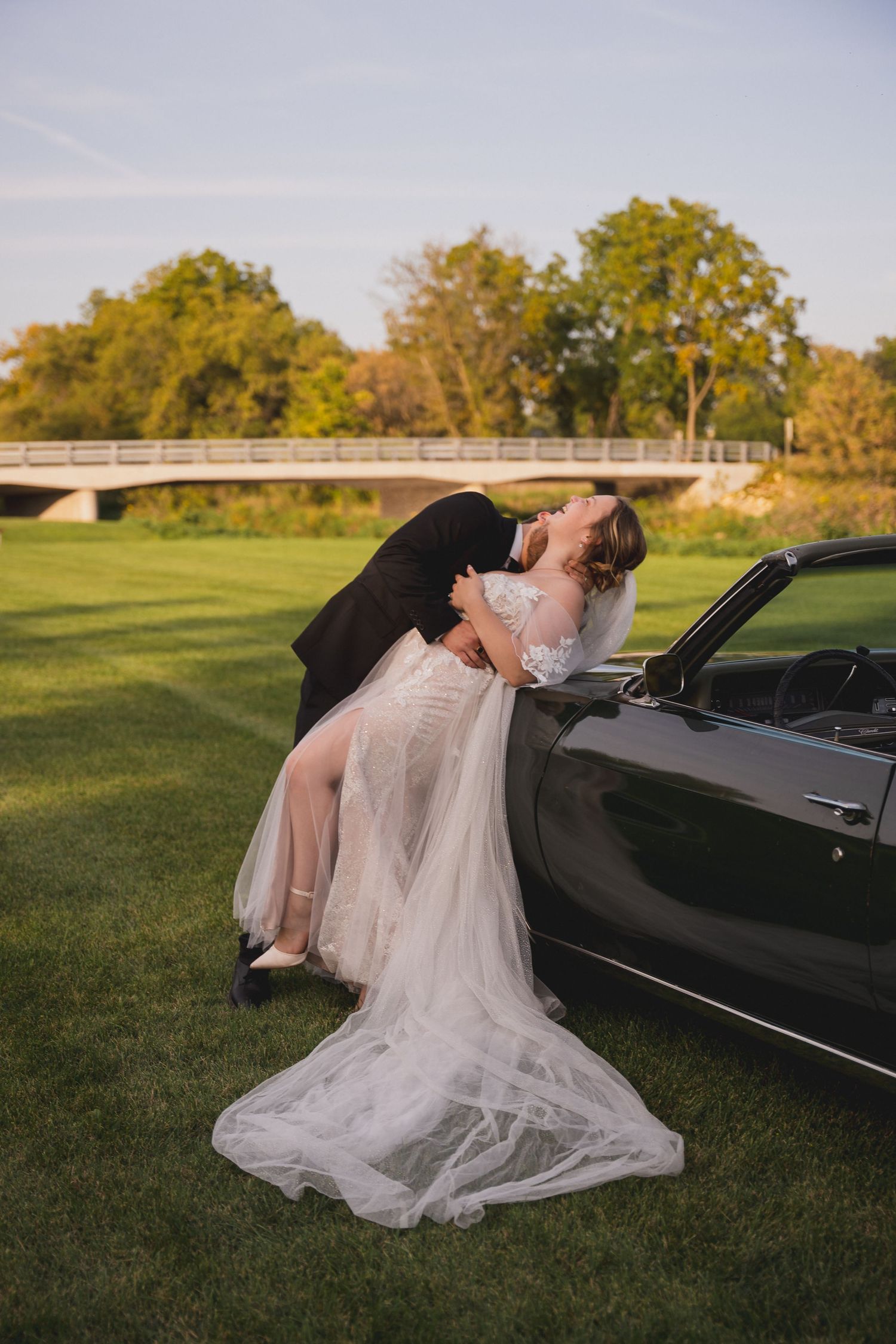 Bride in flowing white dress poses dramatically against classic black car on grassy field.