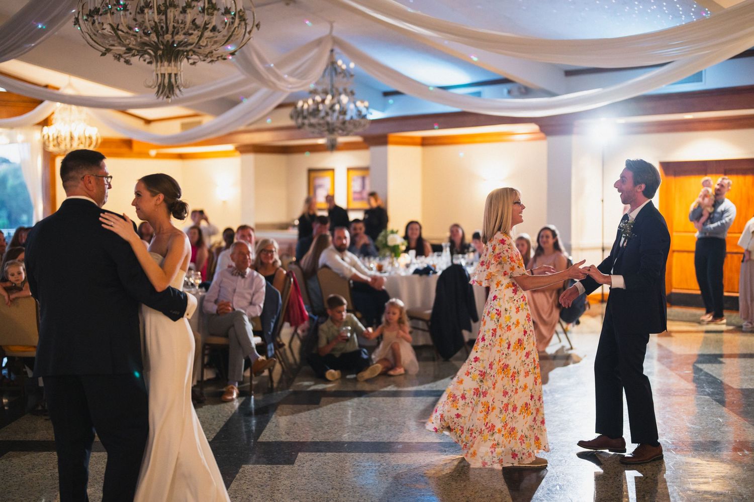 Bride and groom share first dance while guests applaud in decorated ballroom.
