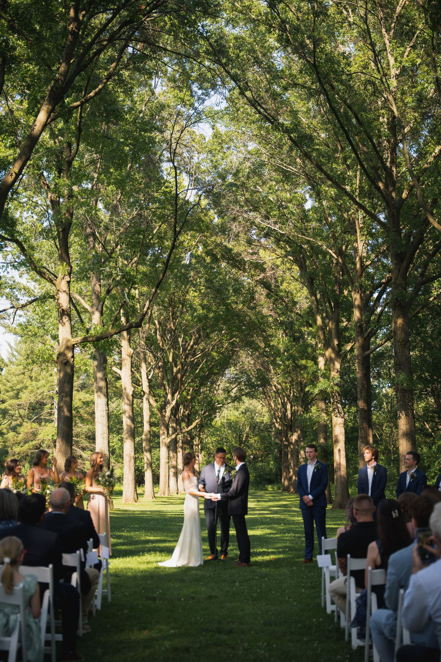 Outdoor wedding ceremony under tall trees with guests seated in white chairs.