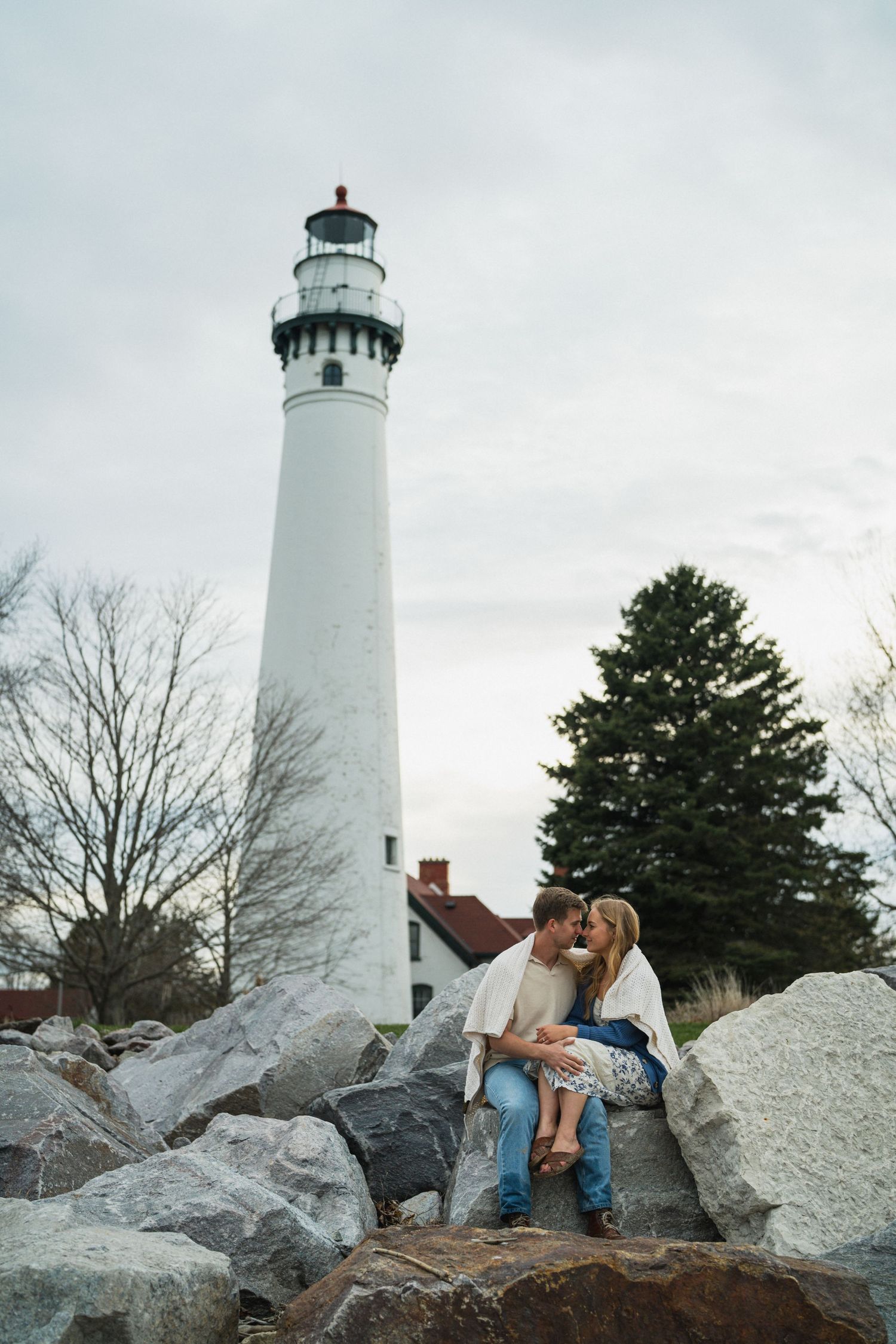 Couple sitting on rocks kissing in front of a tall white lighthouse on a cloudy day.