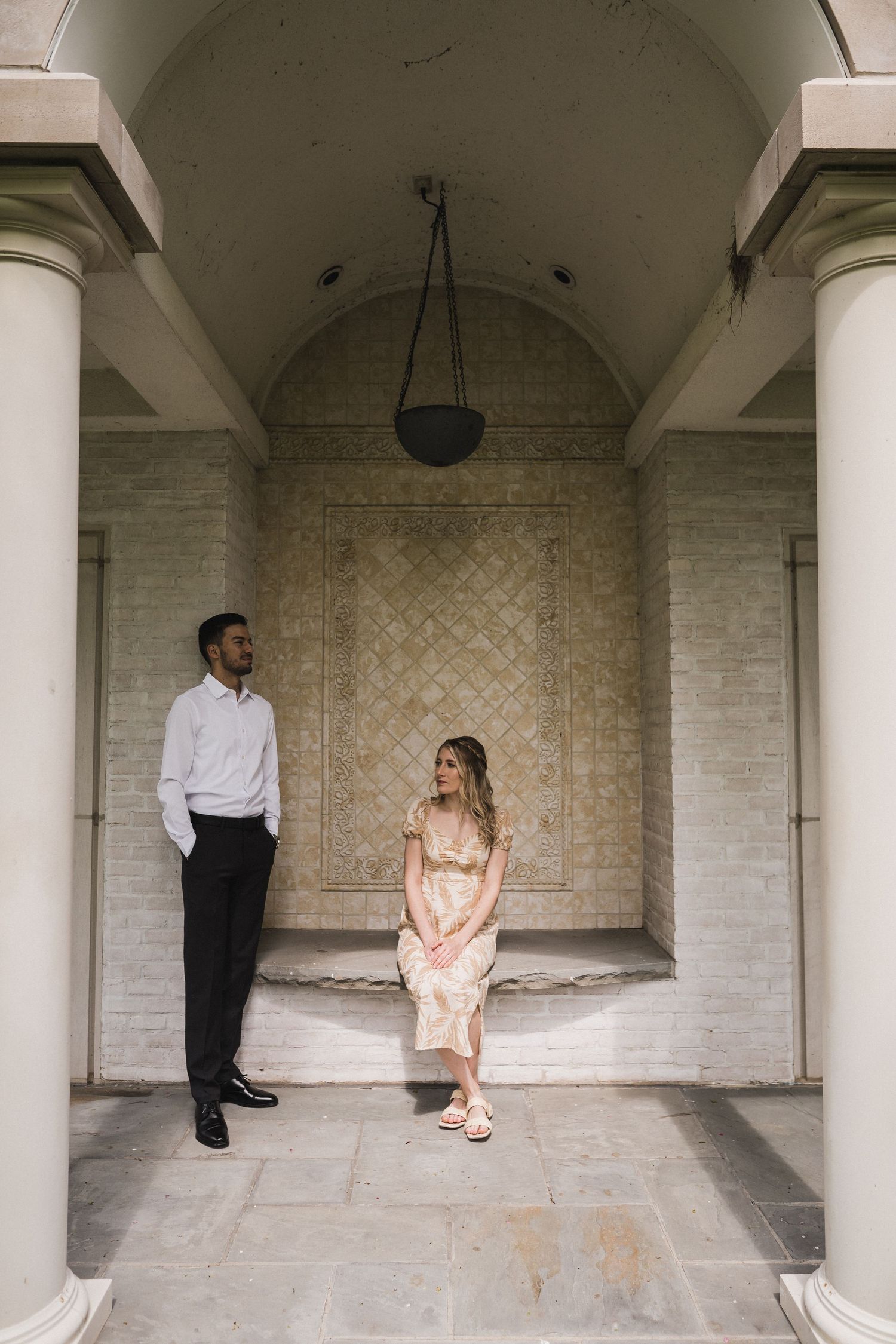 A couple sitting apart under an arched colonnade with white brick walls.