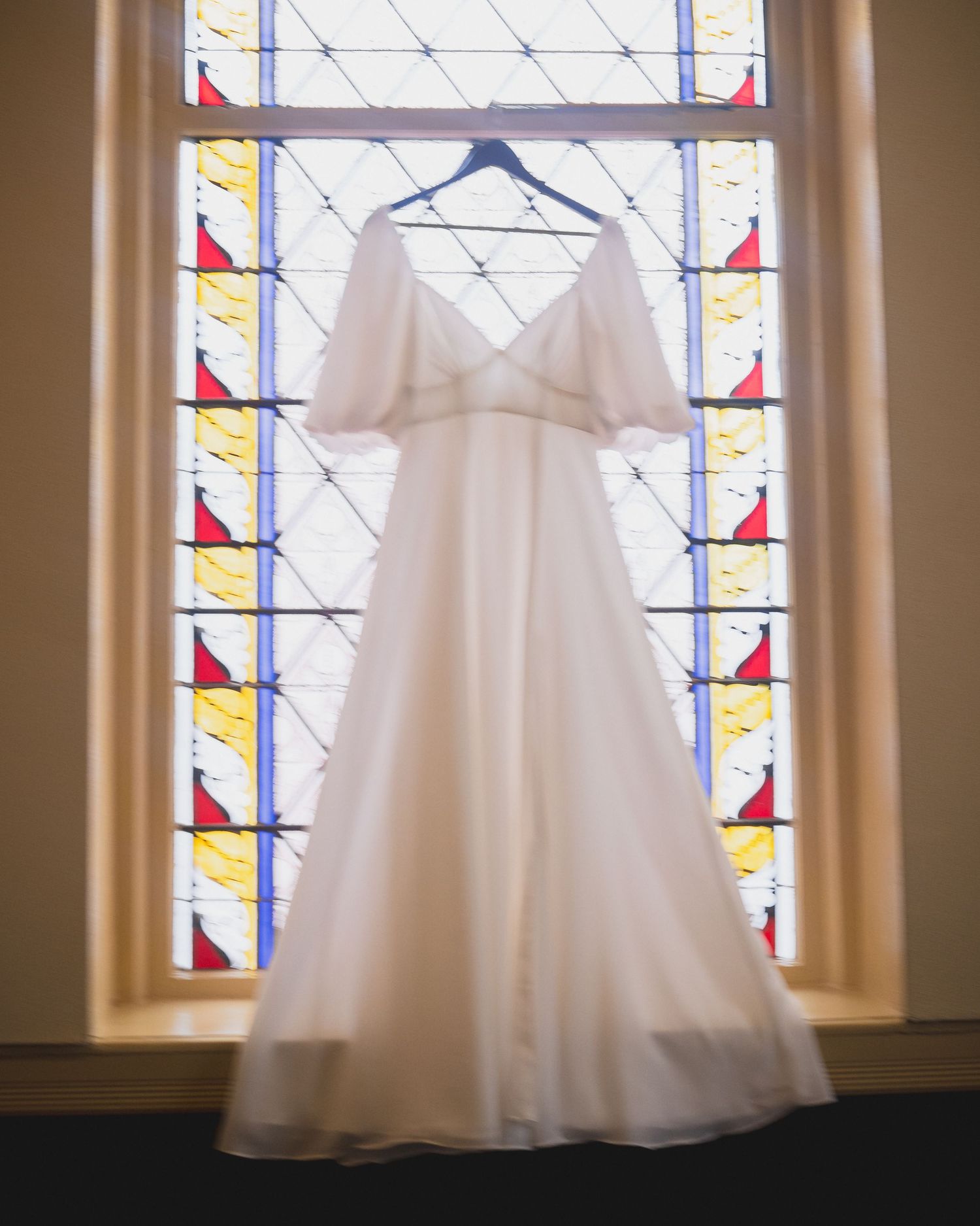 A white wedding dress hanging in front of a stained glass window with geometric patterns.