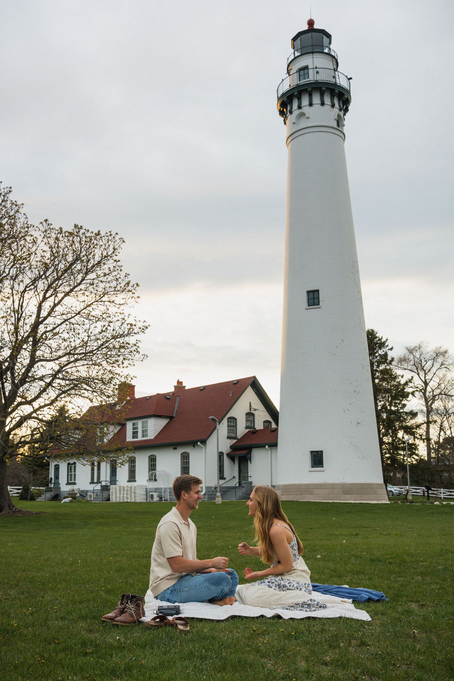 Couple sitting on blanket having picnic on grass beside tall white lighthouse and historic keeper house.