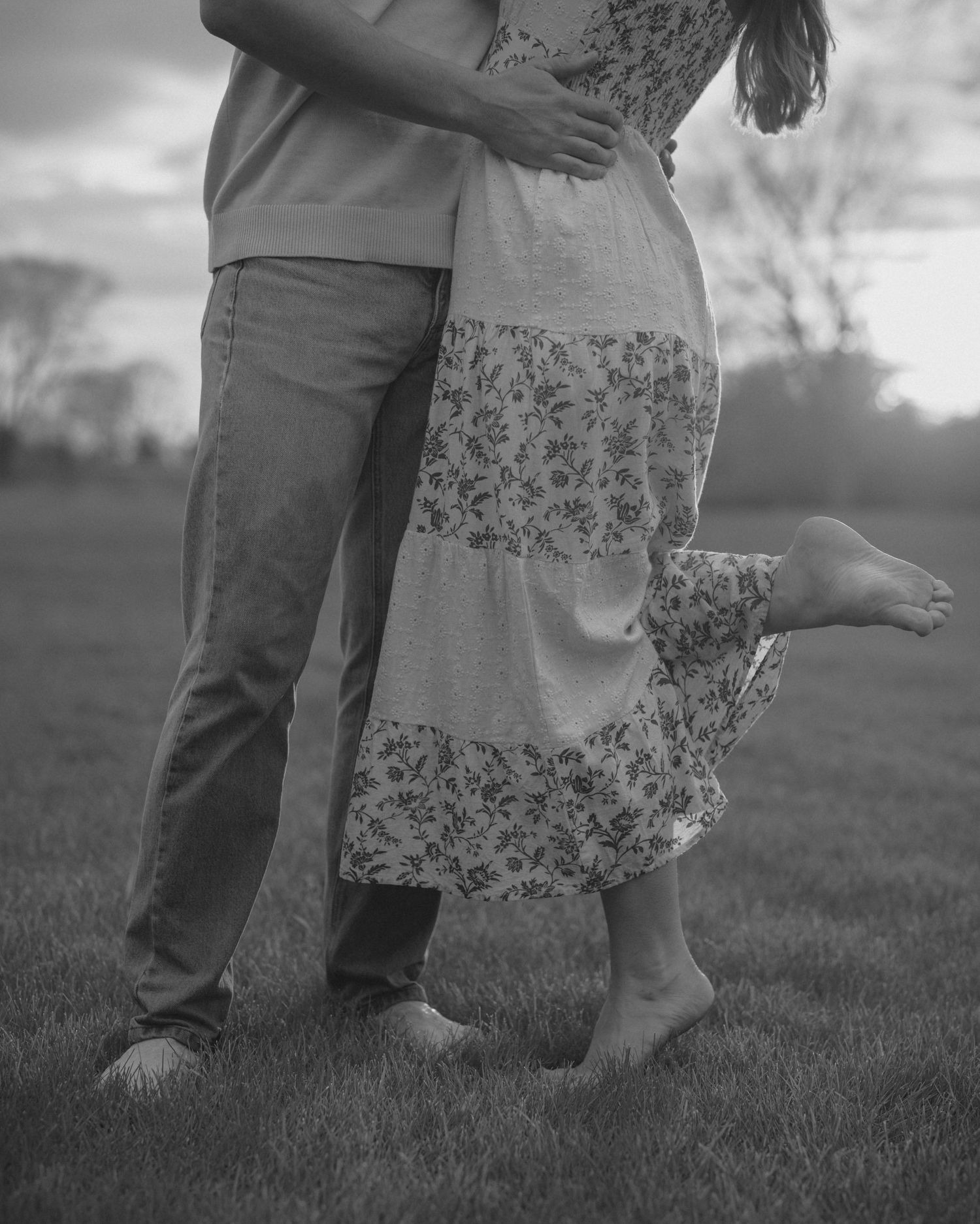 Black and white photo of couple embracing barefoot on grass, woman lifting foot in floral dress.
