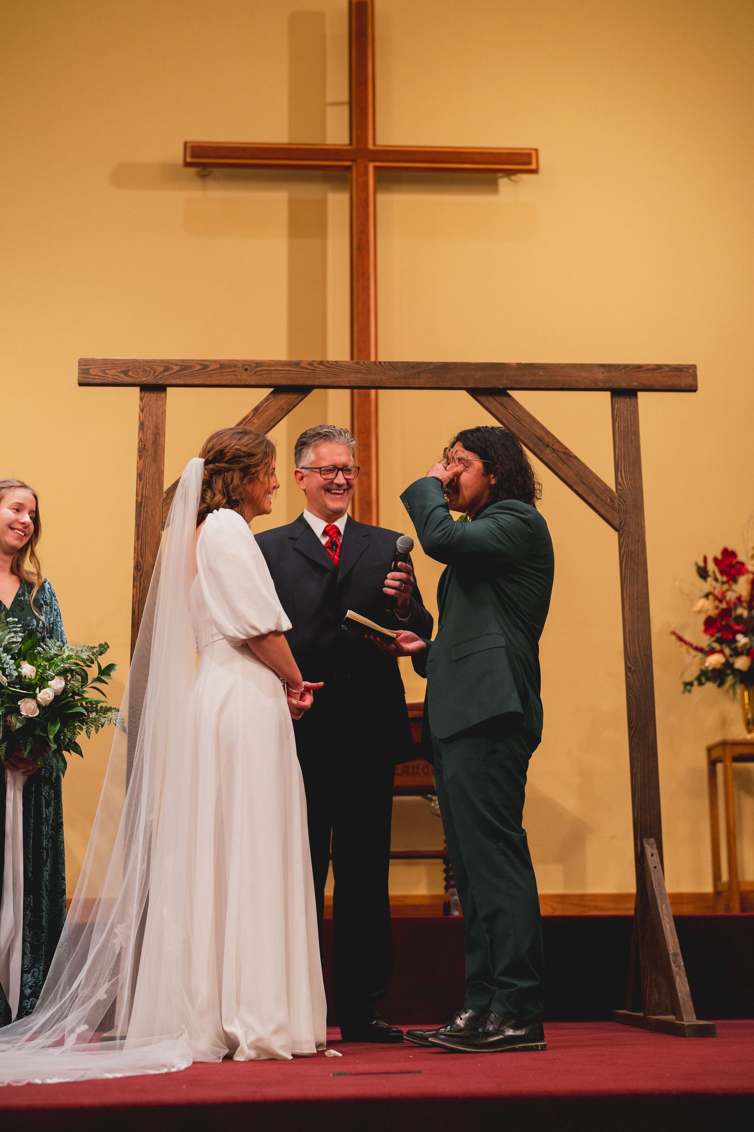 A wedding ceremony takes place under a wooden cross structure with the couple and officiant on a red carpet.