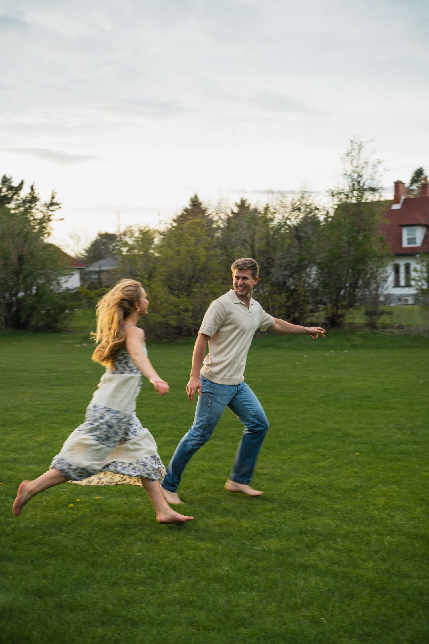 Joyful couple running barefoot on a green lawn with trees and a red-roofed house in background.