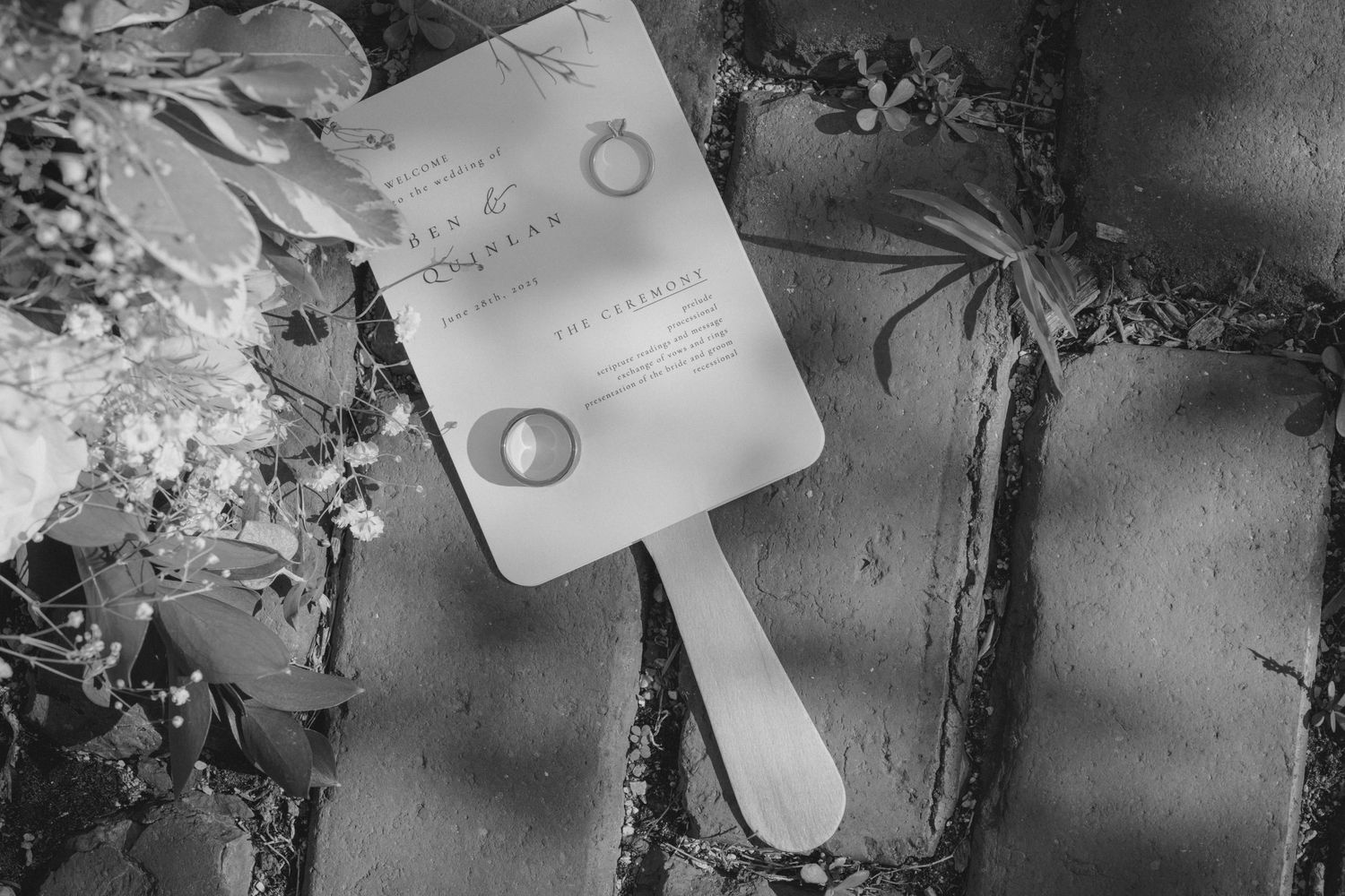Black and white photo of wedding rings and program fan on stone pavers with flowers.