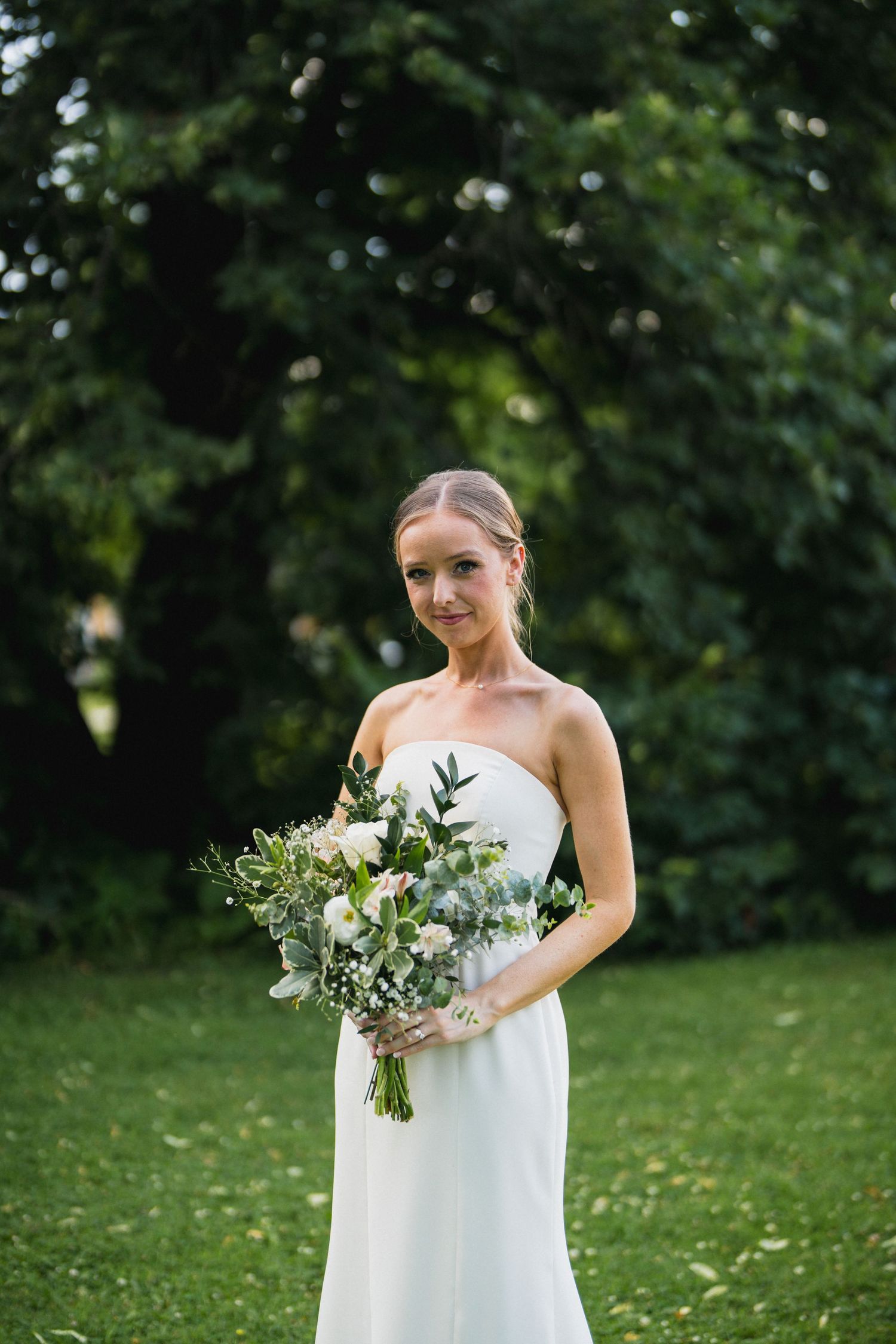 Elegant bride in a strapless white gown holding a white and green bouquet while posing in a garden.
