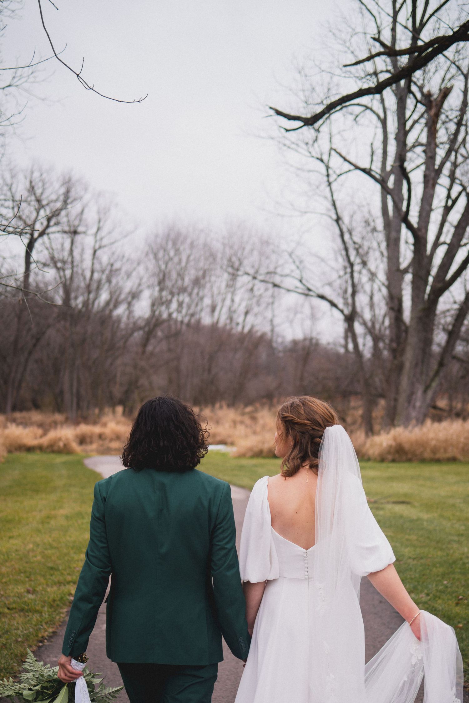 A wedding couple walking through an autumn field with bare trees in the background.