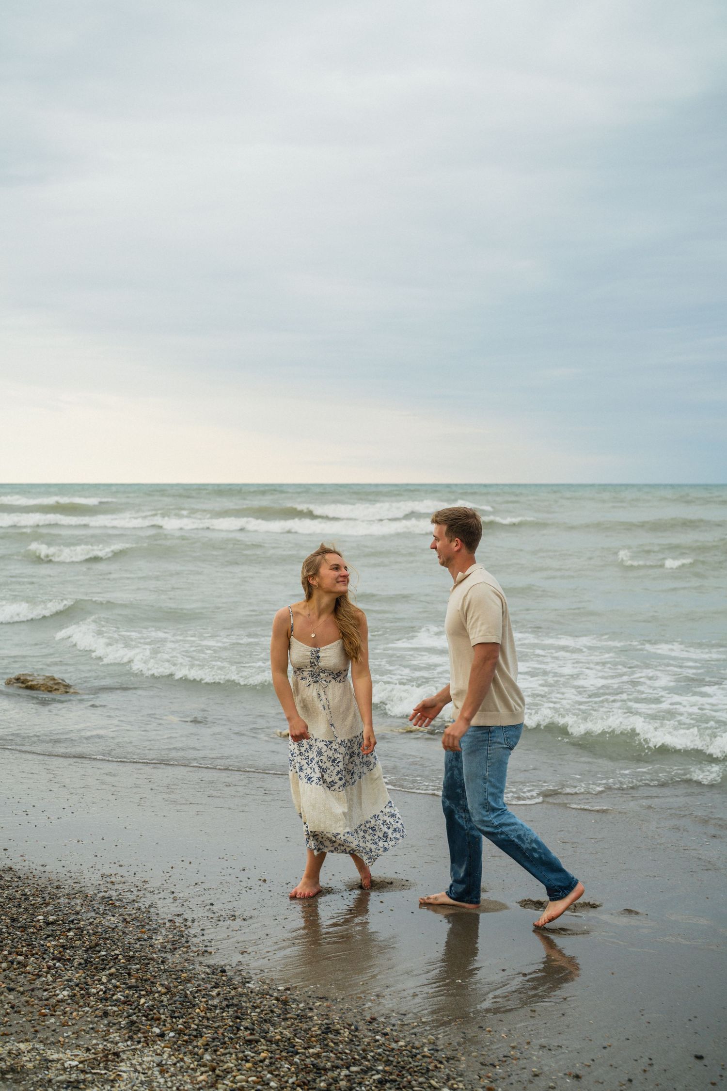 Romantic couple walking barefoot along a rocky beach with ocean waves behind them.