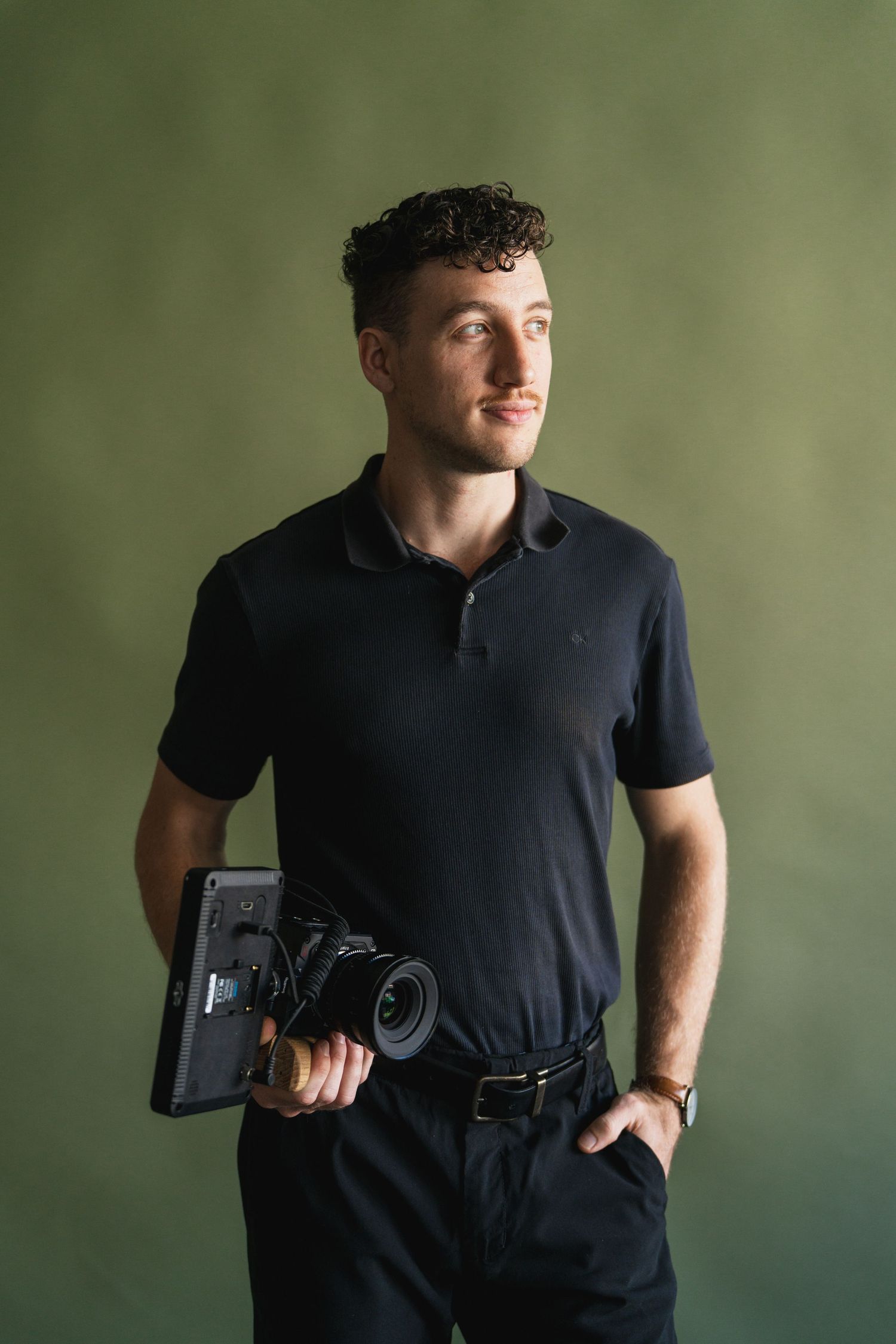 Professional photographer and filmmaker in dark polo shirt poses with camera against green background.