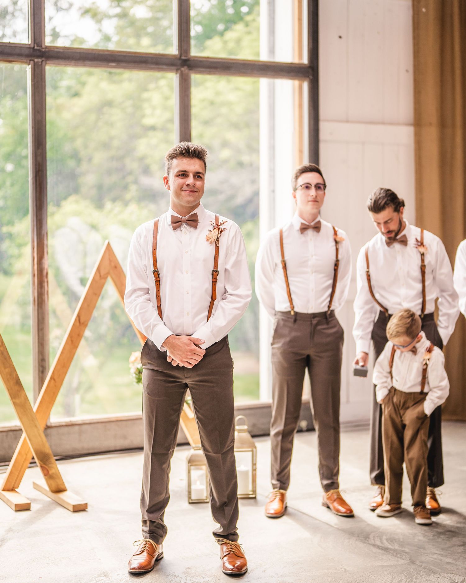 Groomsmen in suspenders and bowties stand in front of large windows with wooden geometric decor at a wedding venue.