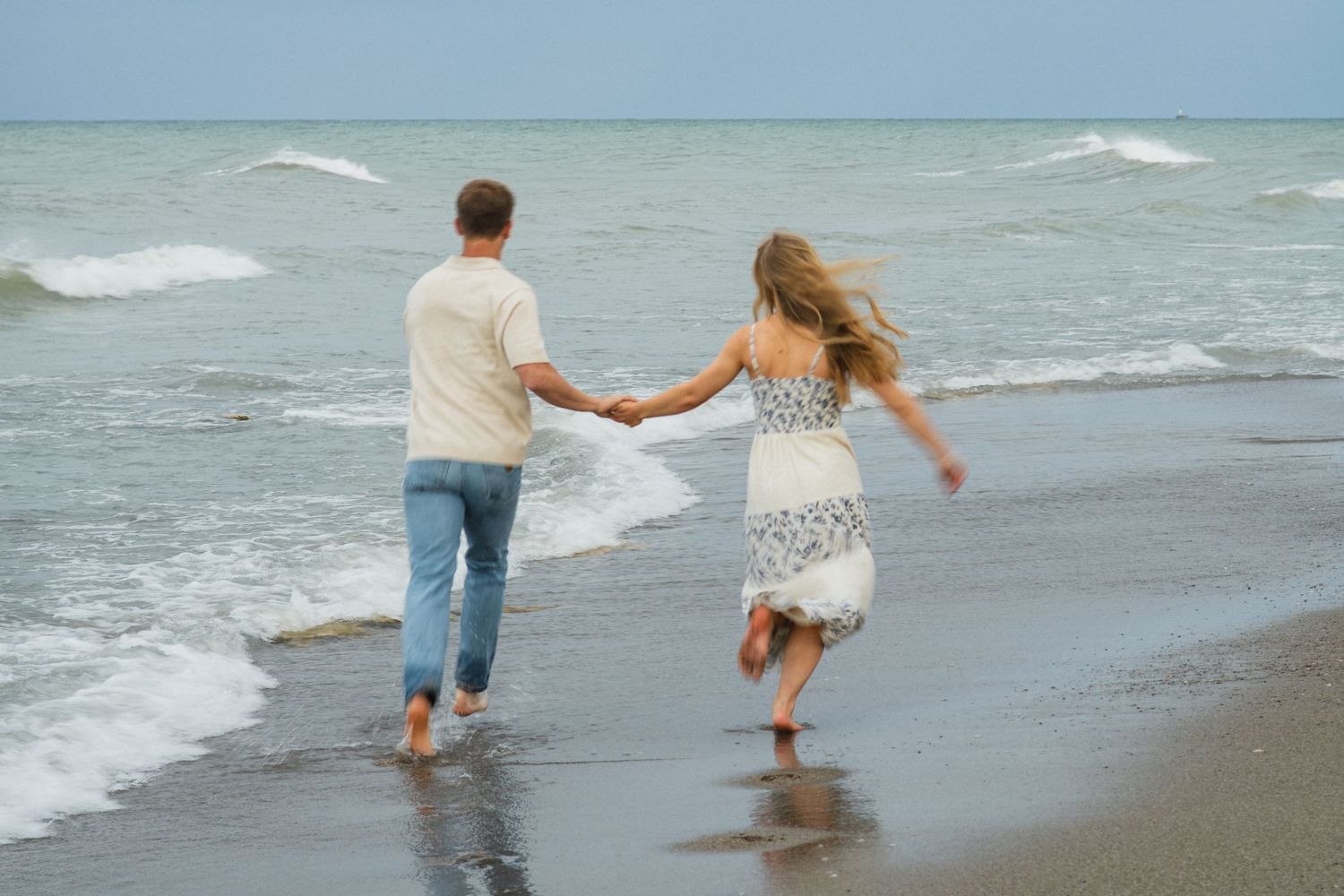 A couple holds hands while running barefoot along a sandy beach with ocean waves in the background.