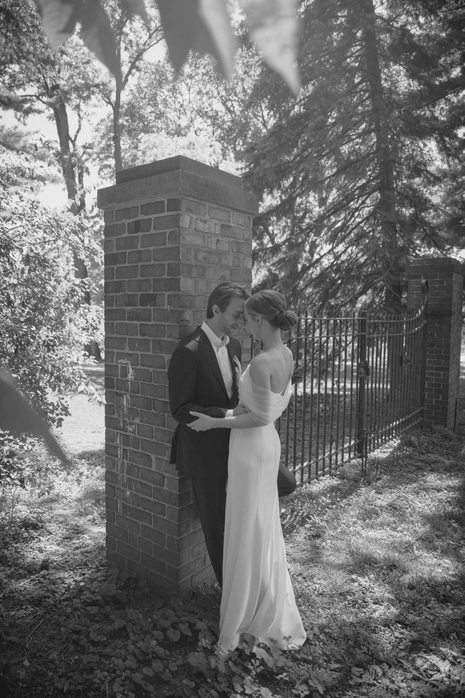 A bride and groom share an intimate moment leaning against a brick pillar in a black and white photo.