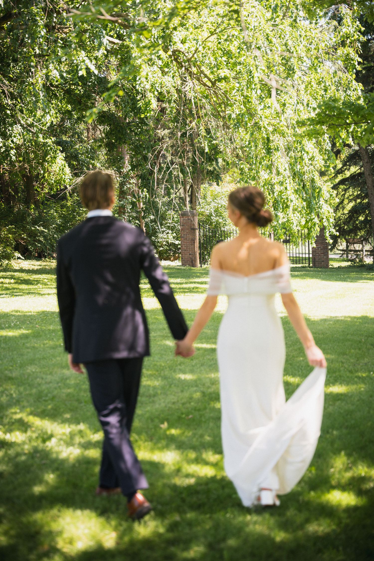 Bride and groom walk hand in hand across lush green lawn, viewed from behind, at outdoor garden wedding.