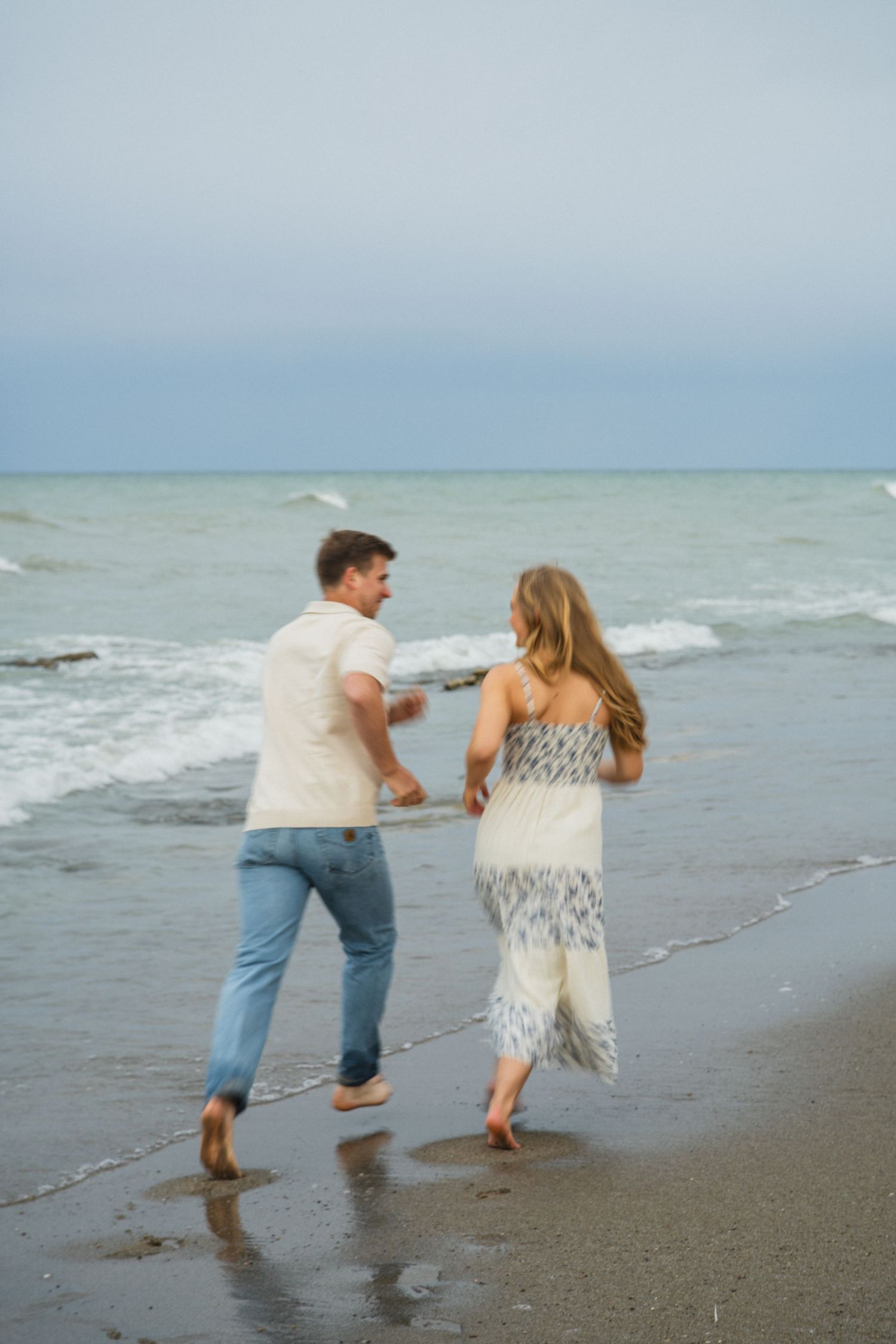 Romantic couple running barefoot along shoreline in casual attire with ocean waves behind them.