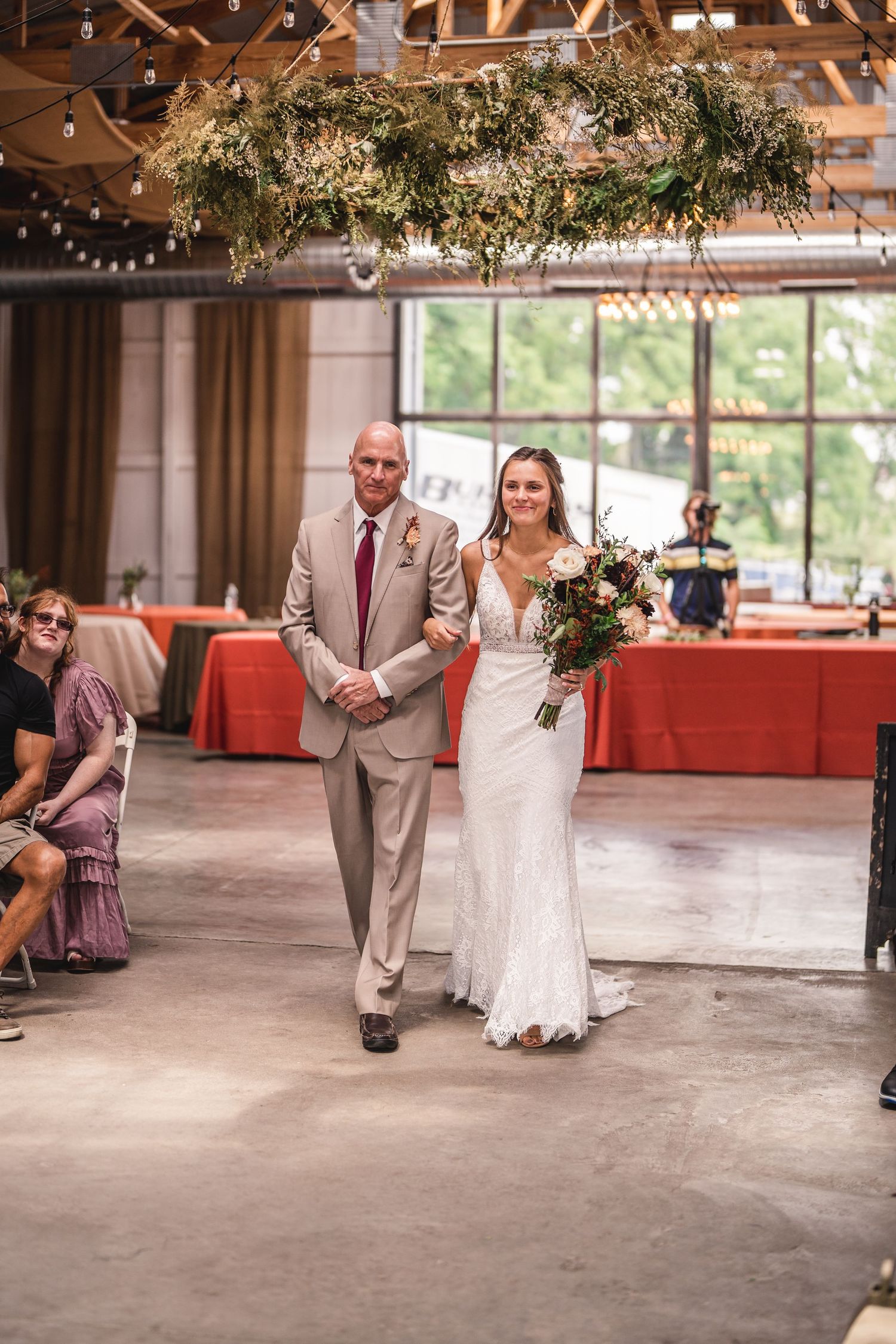 Bride in fitted white gown walks down aisle with escort in industrial venue setting.